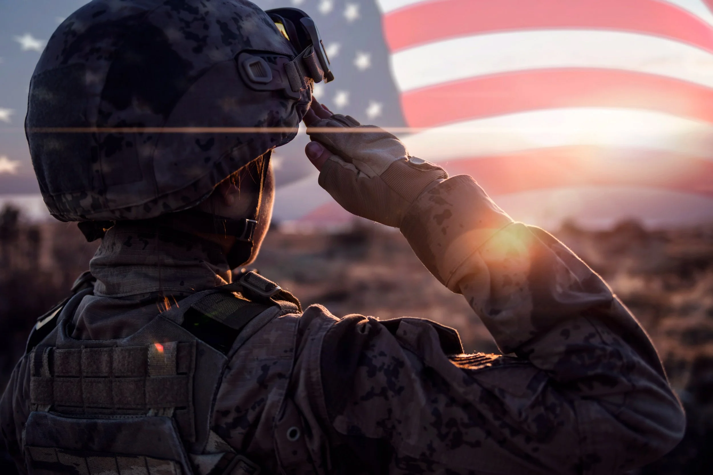A soldier wearing camouflage uniform, helmet, goggles, and gloves salutes with a hand on his forehead during sunset, with an American flag in the background.