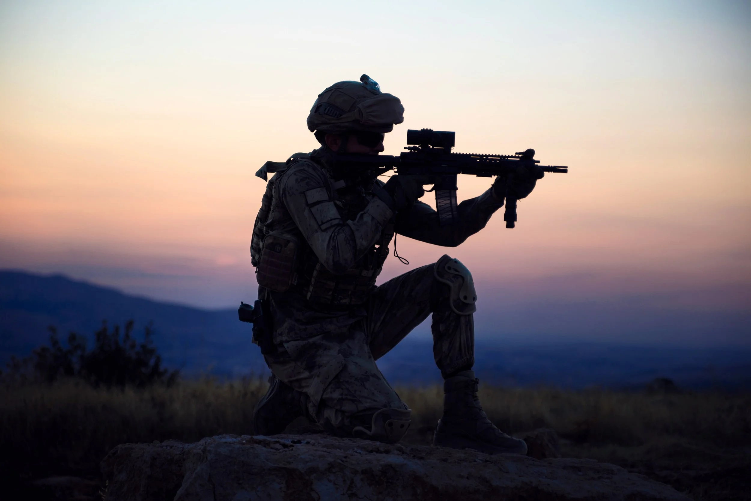 A soldier in camouflage uniform, helmet, and tactical gear kneeling on a rock and aiming a sniper rifle during sunset or dusk.