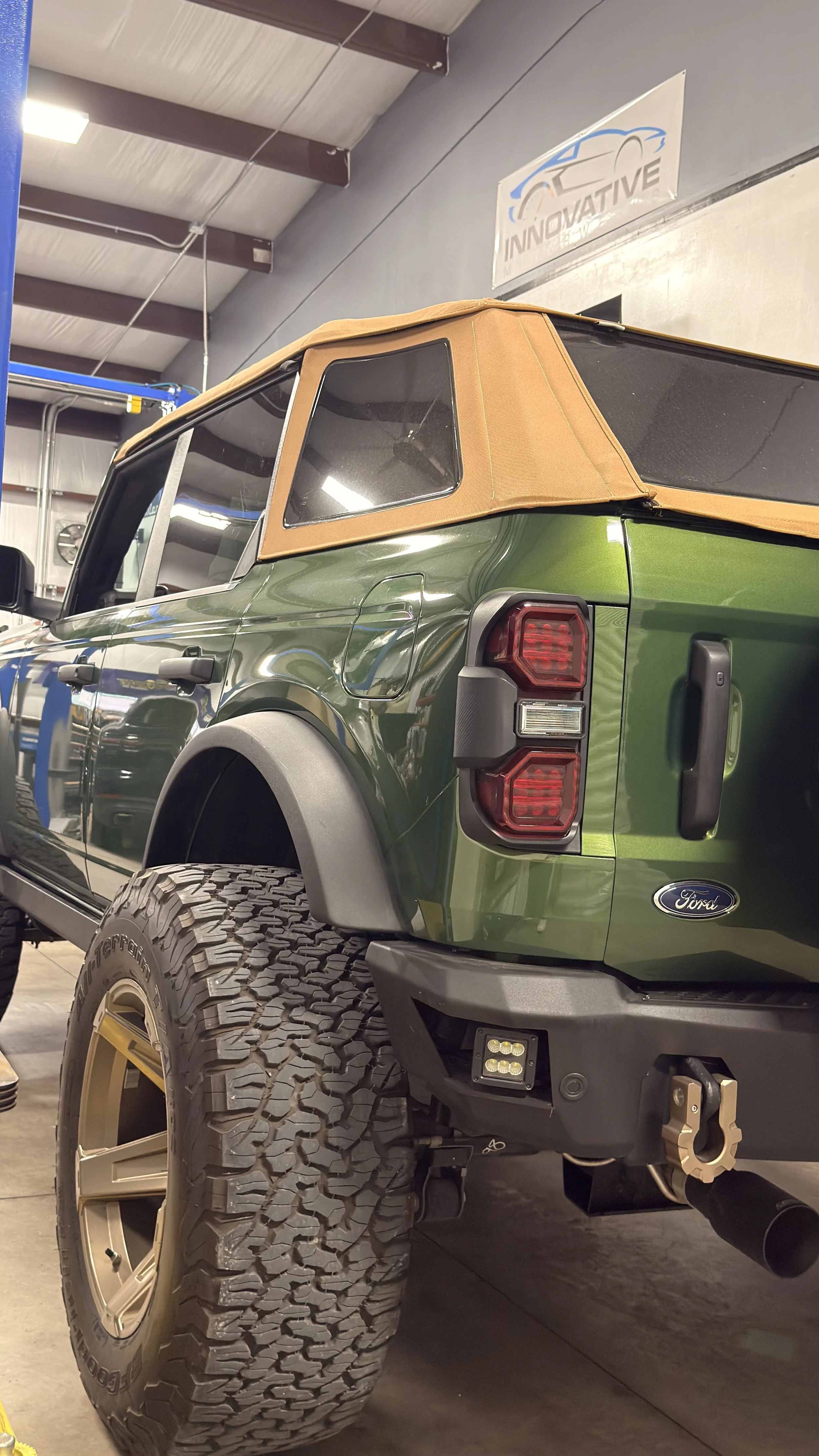 Green Ford off-road vehicle with large tires, tan soft top, and off-road accessories inside a garage or showroom near a sign that reads "Innovative".