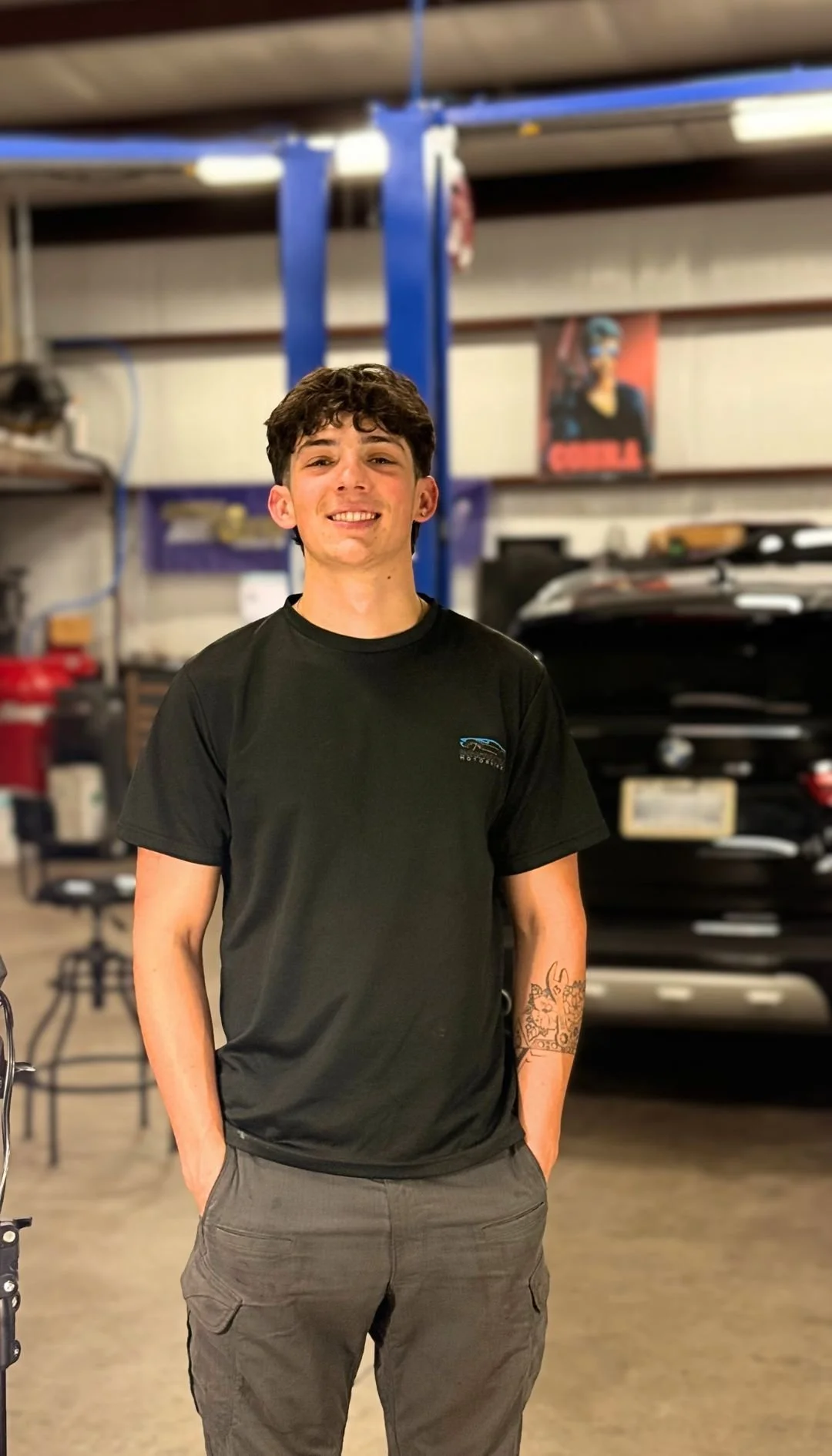 A young man with curly dark hair smiling, standing in a garage with a car behind him, wearing a black t-shirt and gray cargo pants.