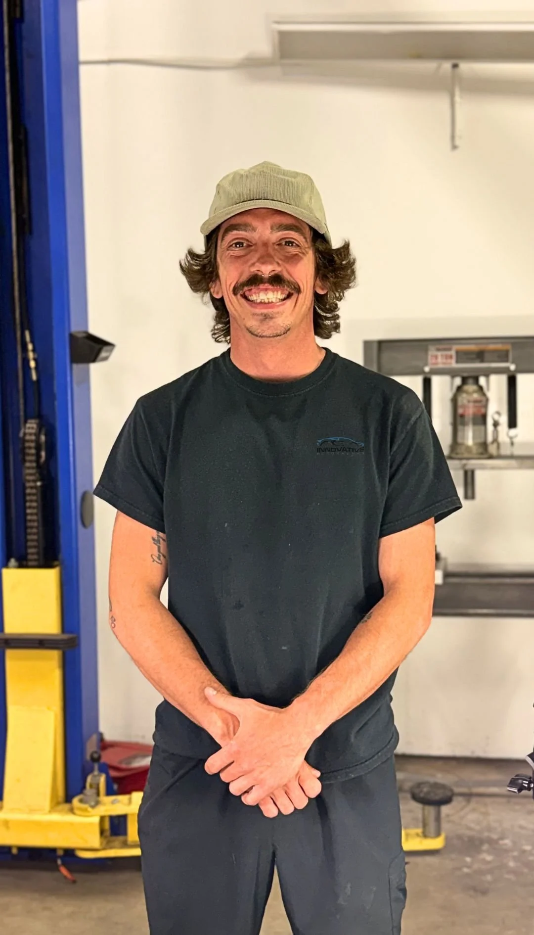 A man with a beard and mustache, wearing a beige cap and black T-shirt, standing in a garage or workshop with a yellow and blue vehicle lift and some equipment in the background.