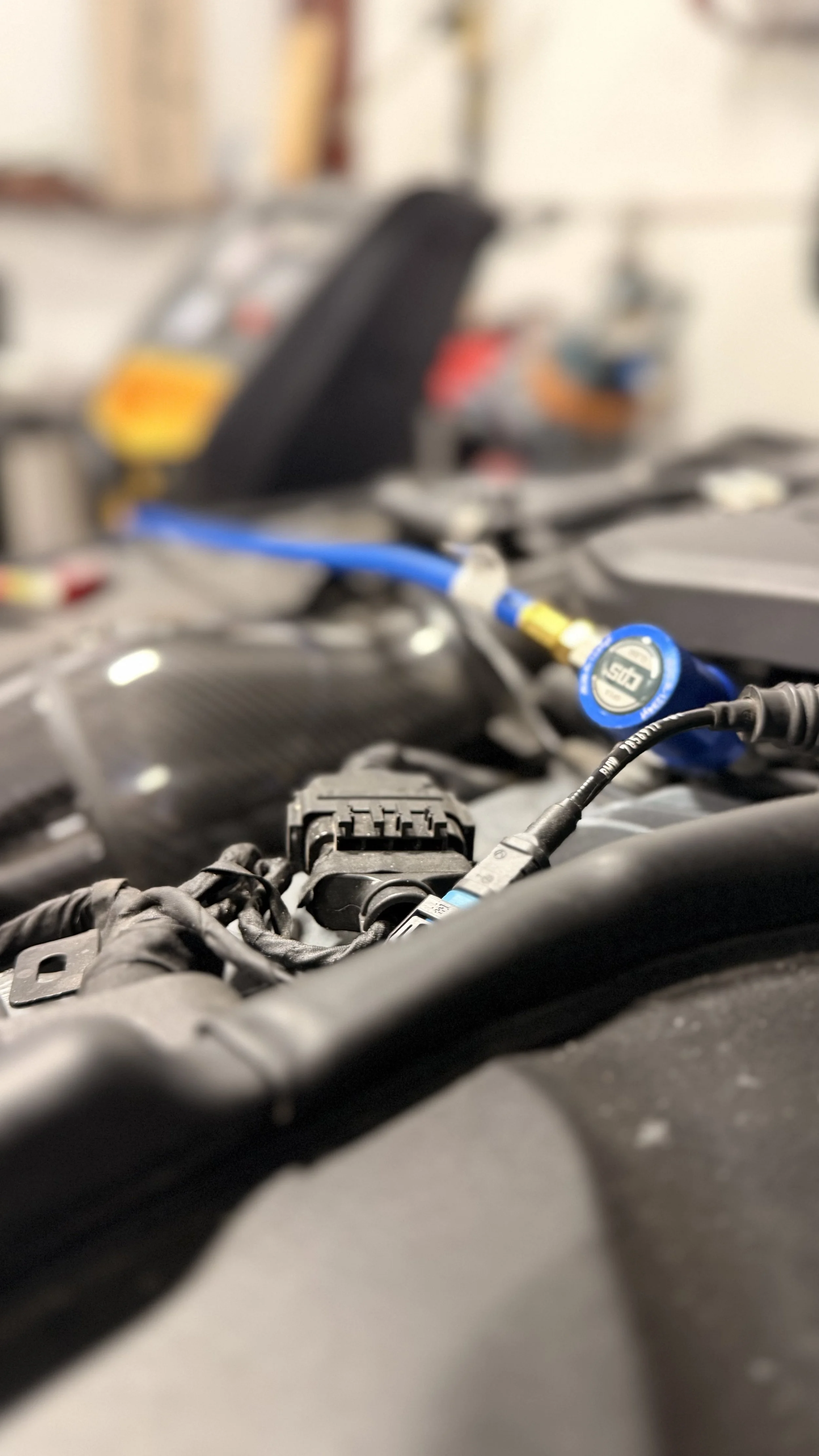 Close-up of car engine components with wires and connectors, and a blurred background of tools and equipment.