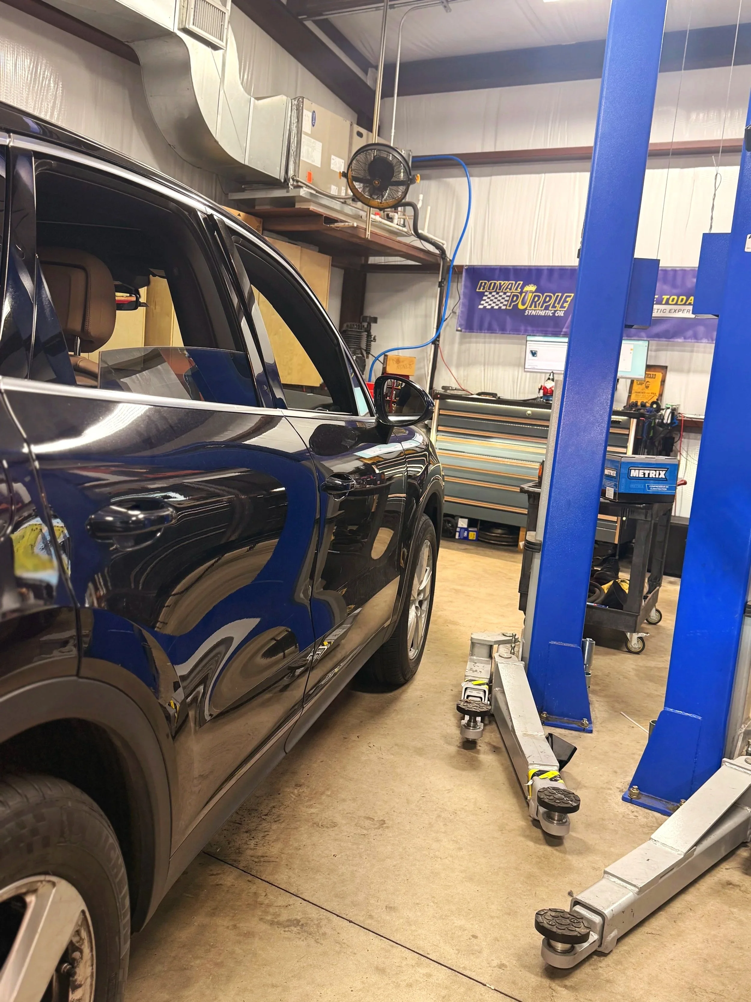 A black SUV vehicle inside an automotive repair shop, lifted on a hydraulic lift. The shop has tools and equipment, including a workbench, drawers, and banners for Royal Purple synthetic oil.