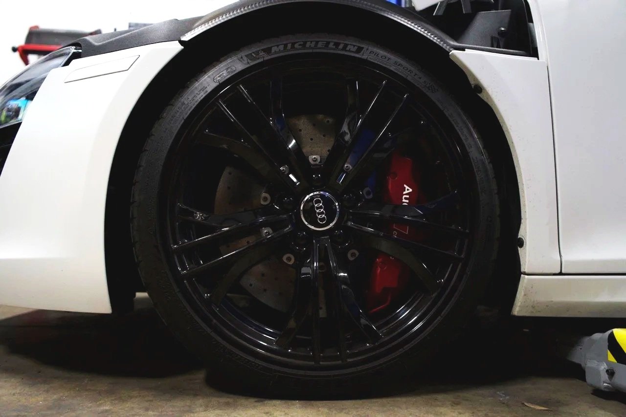 Close-up of a white luxury sports car's front left wheel, featuring a black rim with the Audi logo, red brake caliper, and Michelin Pilot Sport tire, parked indoors.
