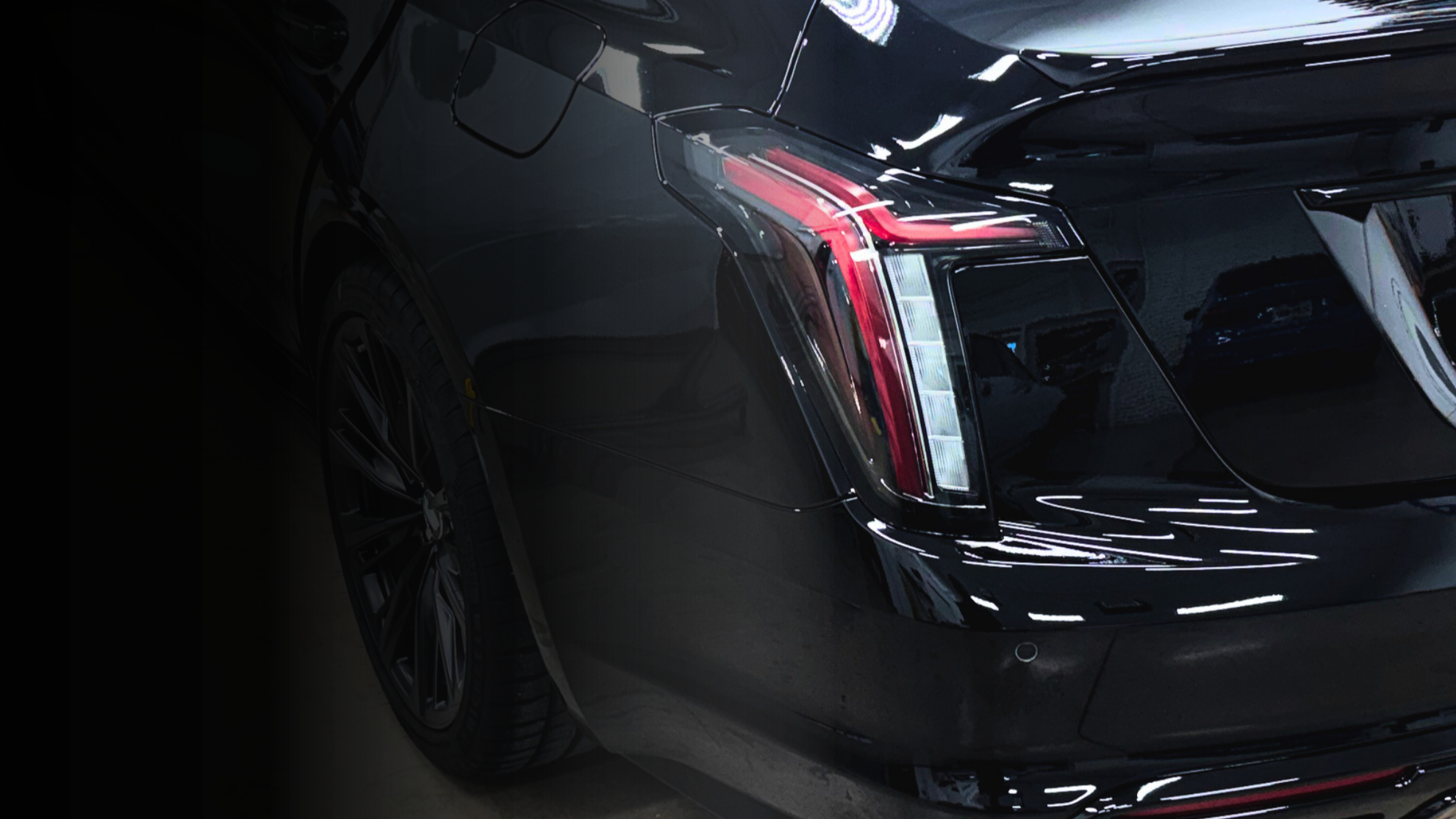 Close-up of the rear end of a black luxury car, showing the left taillight and part of the rear bumper, with reflective surfaces and sleek design.