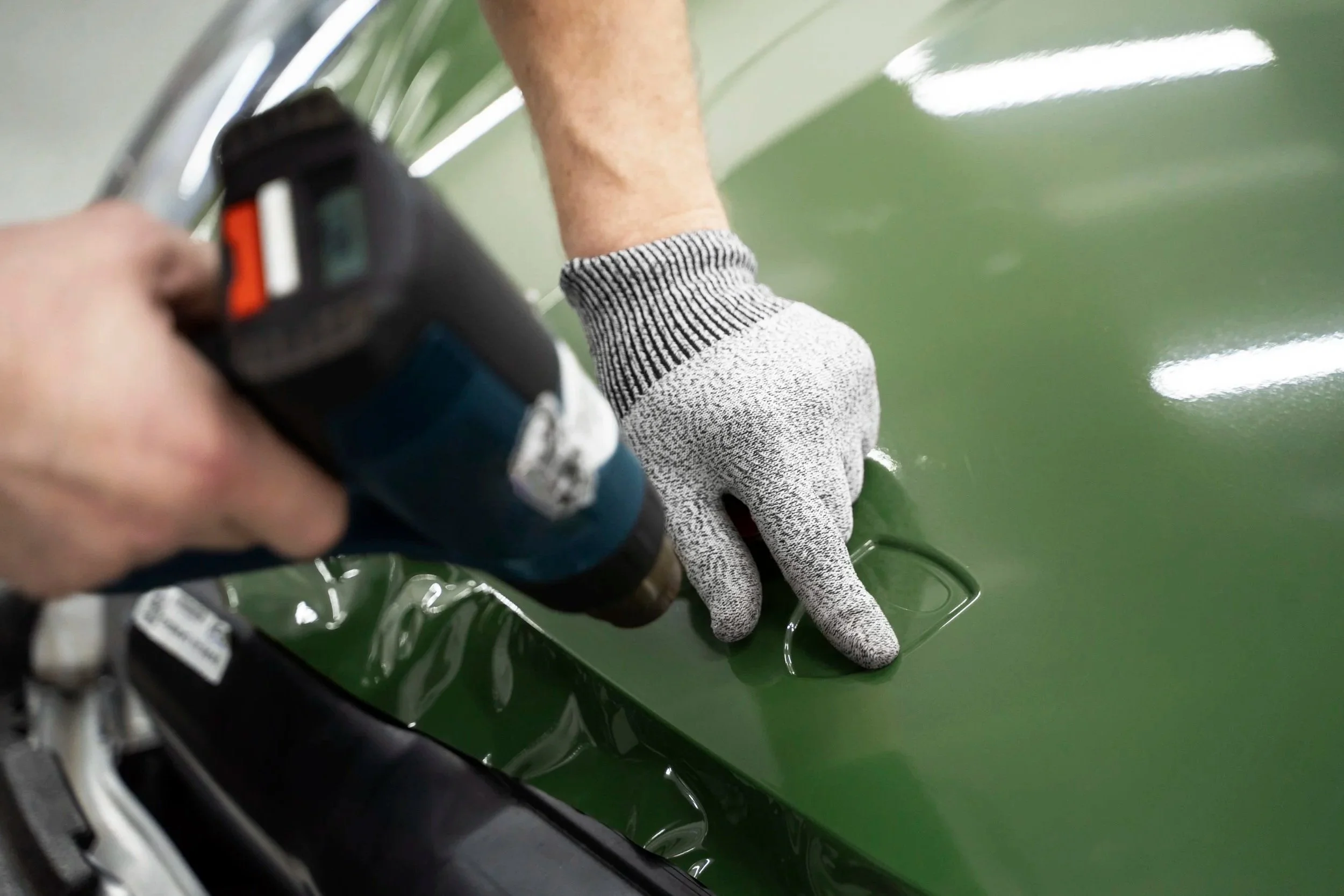 A person wearing gray gloves is working on a green car, using a power tool to polish or buff the surface.