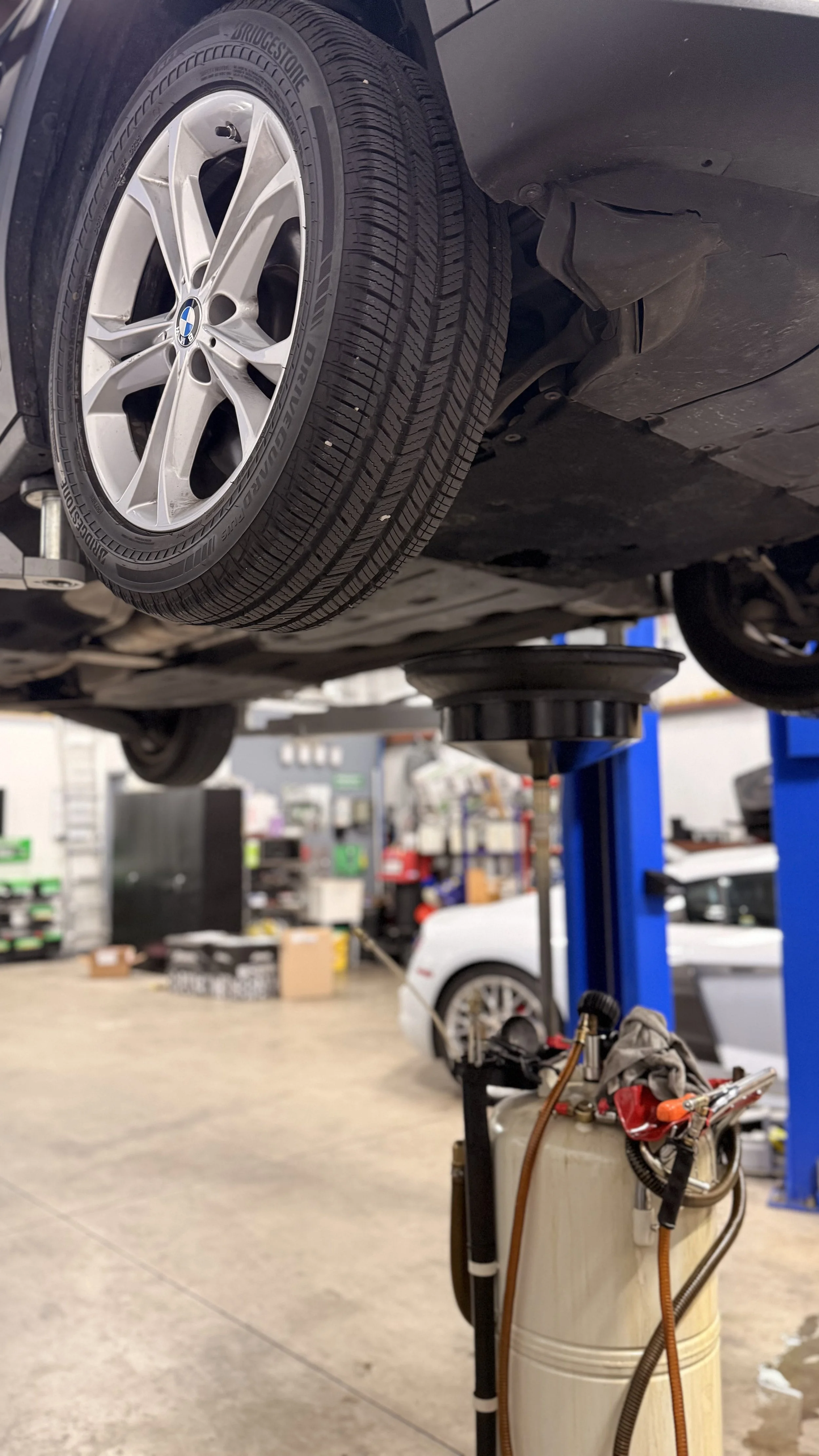 Underbody view of a car on a lift in an auto repair shop, showing a tire with a BMW wheel, mechanic tools, and equipment in the background.