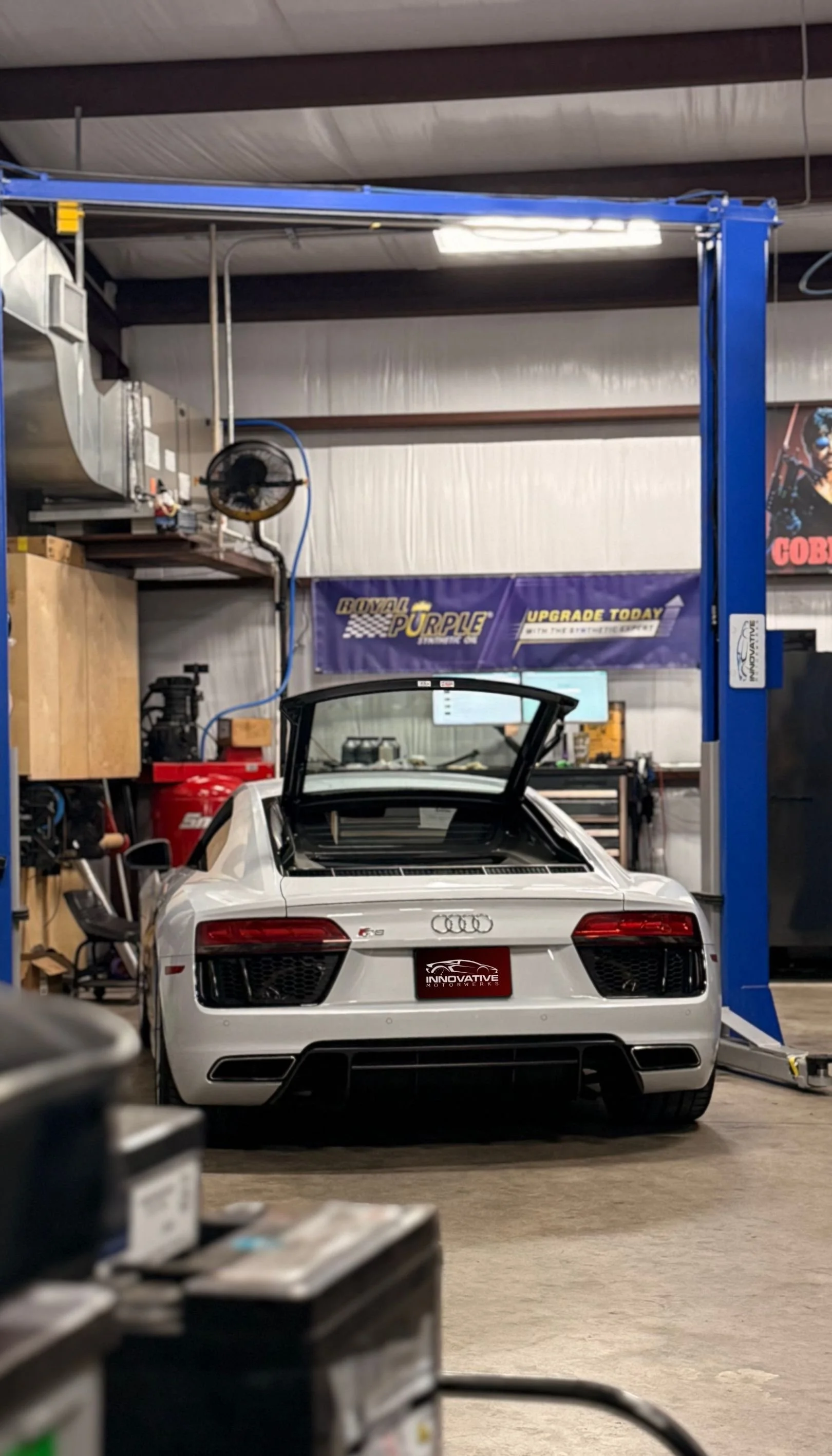 White Audi sports car with rear hatch open inside an automotive workshop or garage.