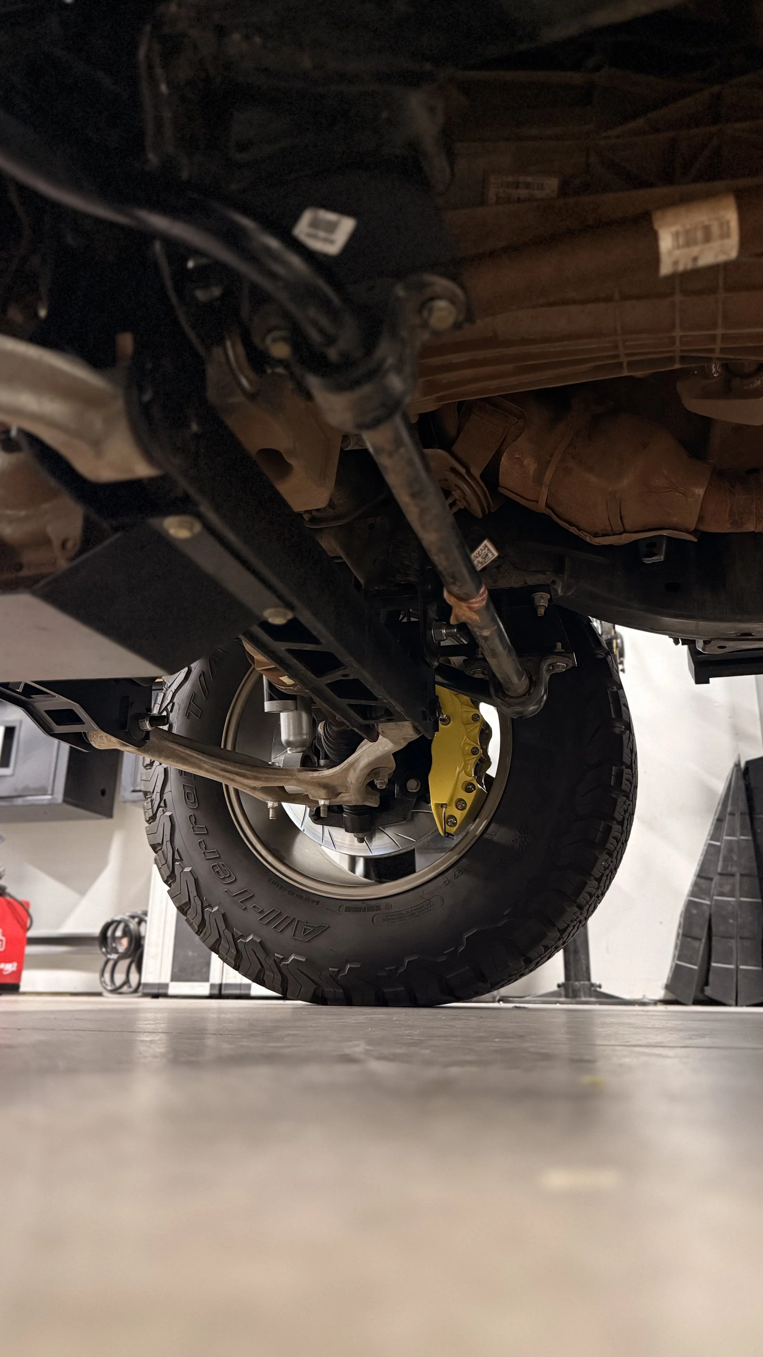 View of an underside of a vehicle showing a large off-road tire, yellow brake caliper, and suspension components in a garage or workshop.