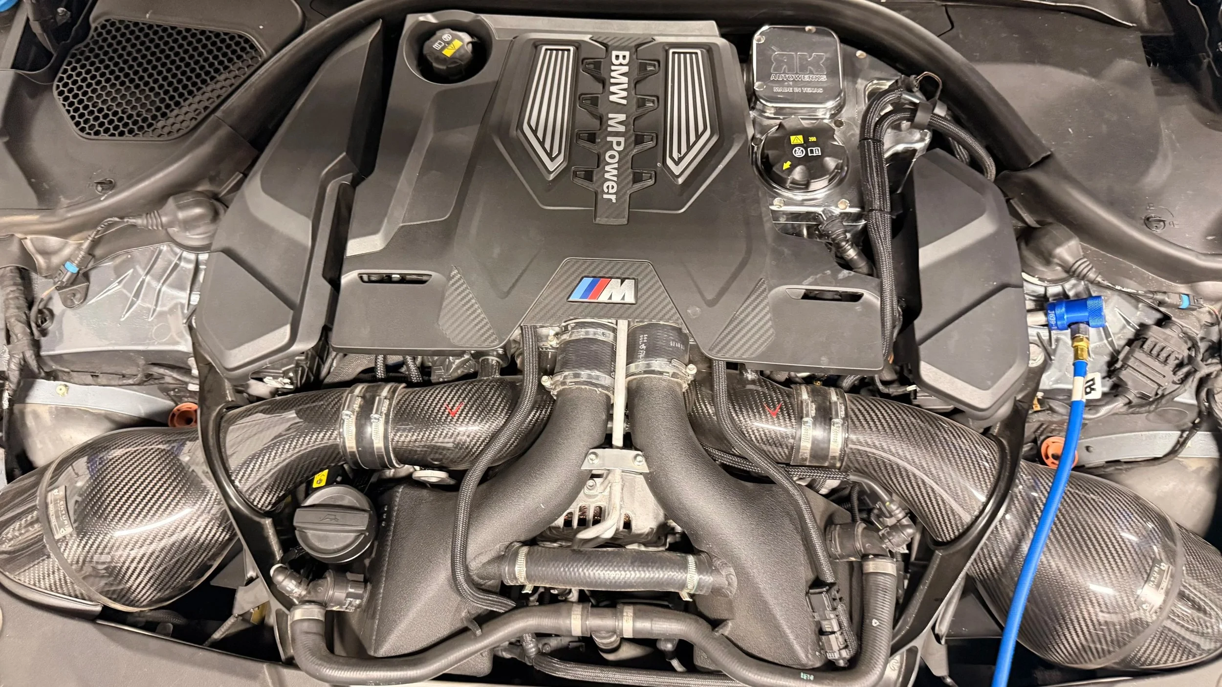A BMW M Power engine with carbon fiber intake pipes, engine cover with BMW M logo, and various interconnected hoses and components.