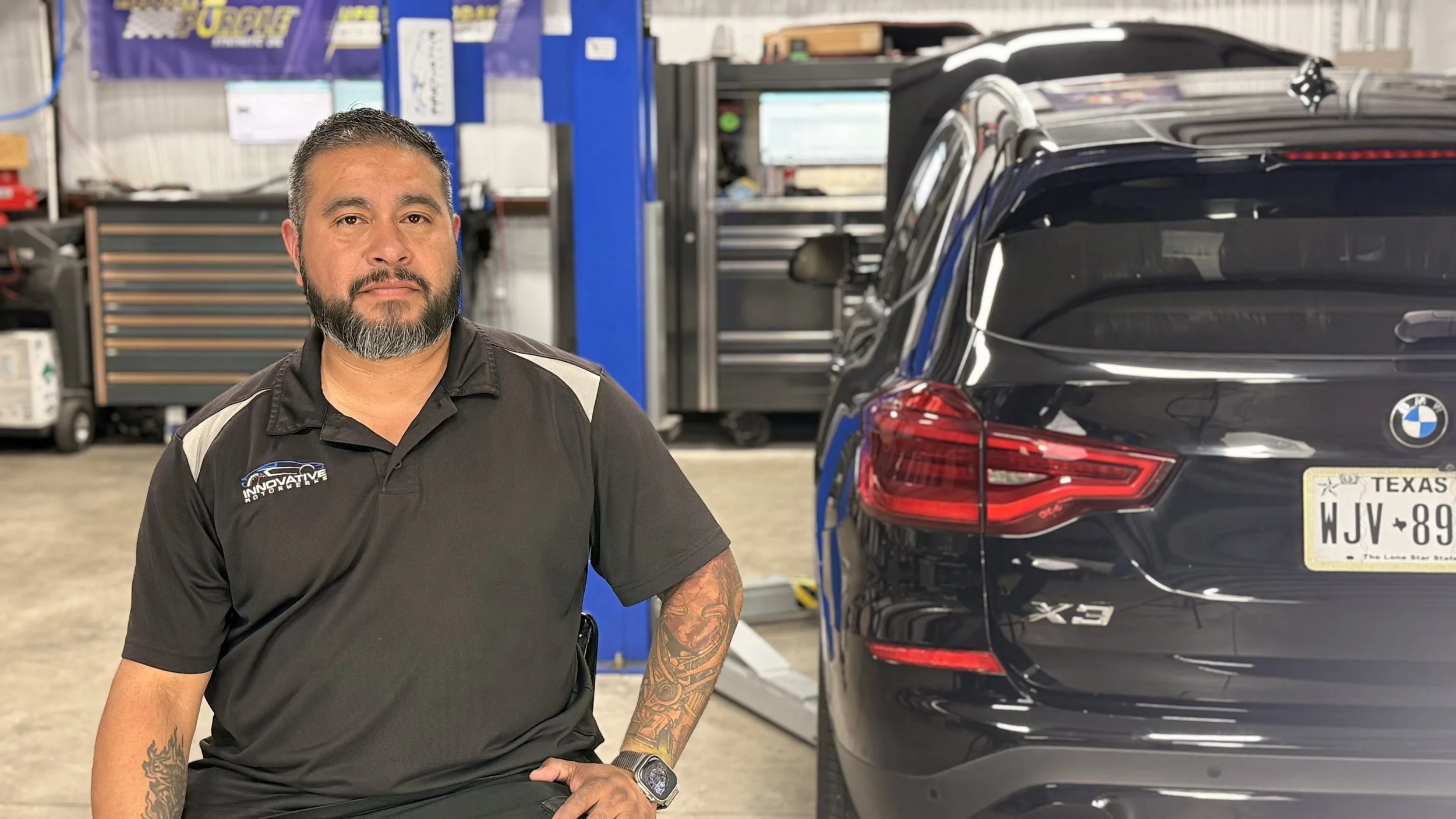 A man with a beard and tattoos on his arms wearing a black work shirt is standing in an auto repair shop next to a black BMW X3 SUV with Texas license plates.