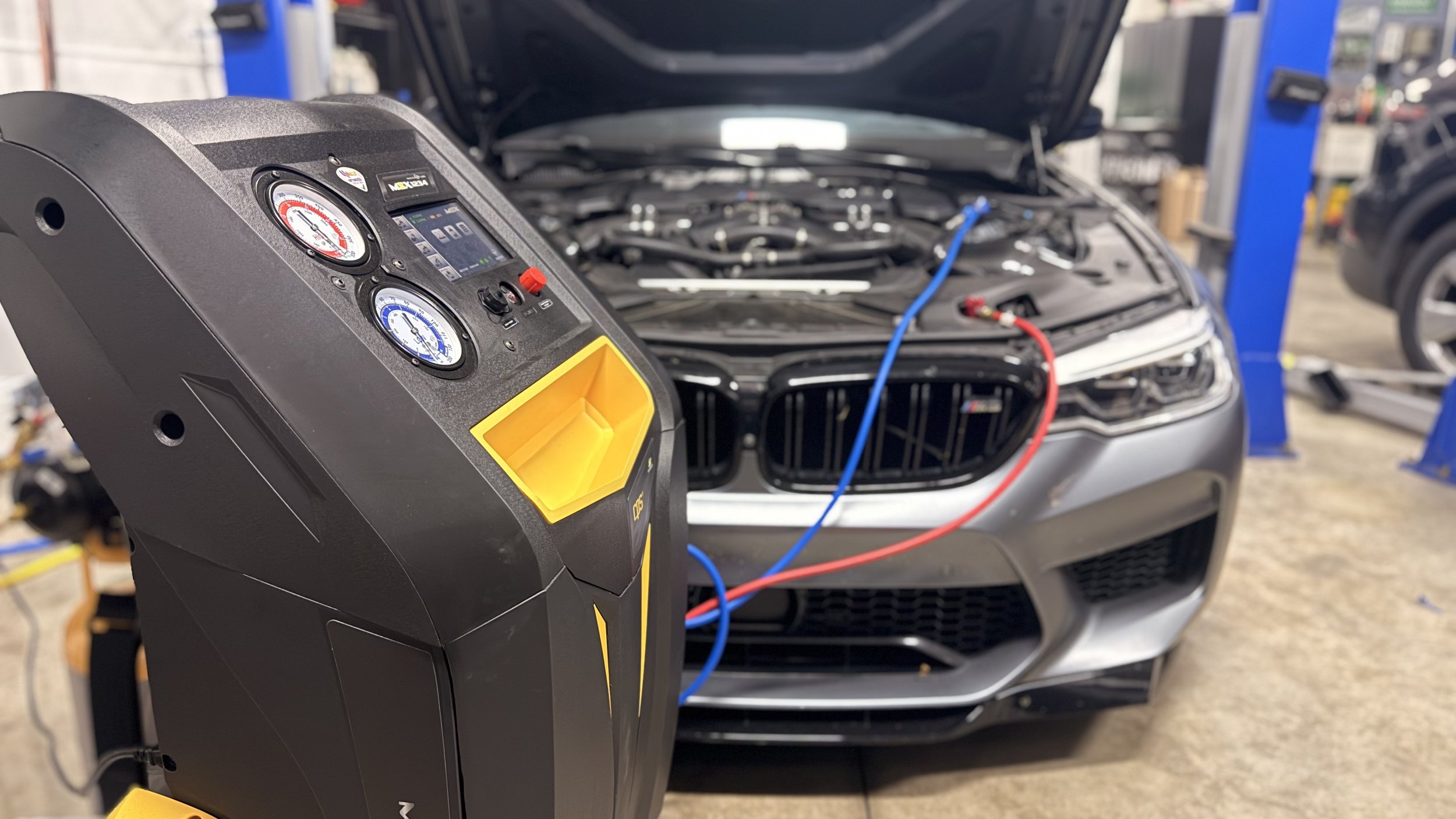 A digital automotive air conditioning service machine with gauges and hoses connected to a car engine in an auto repair shop.