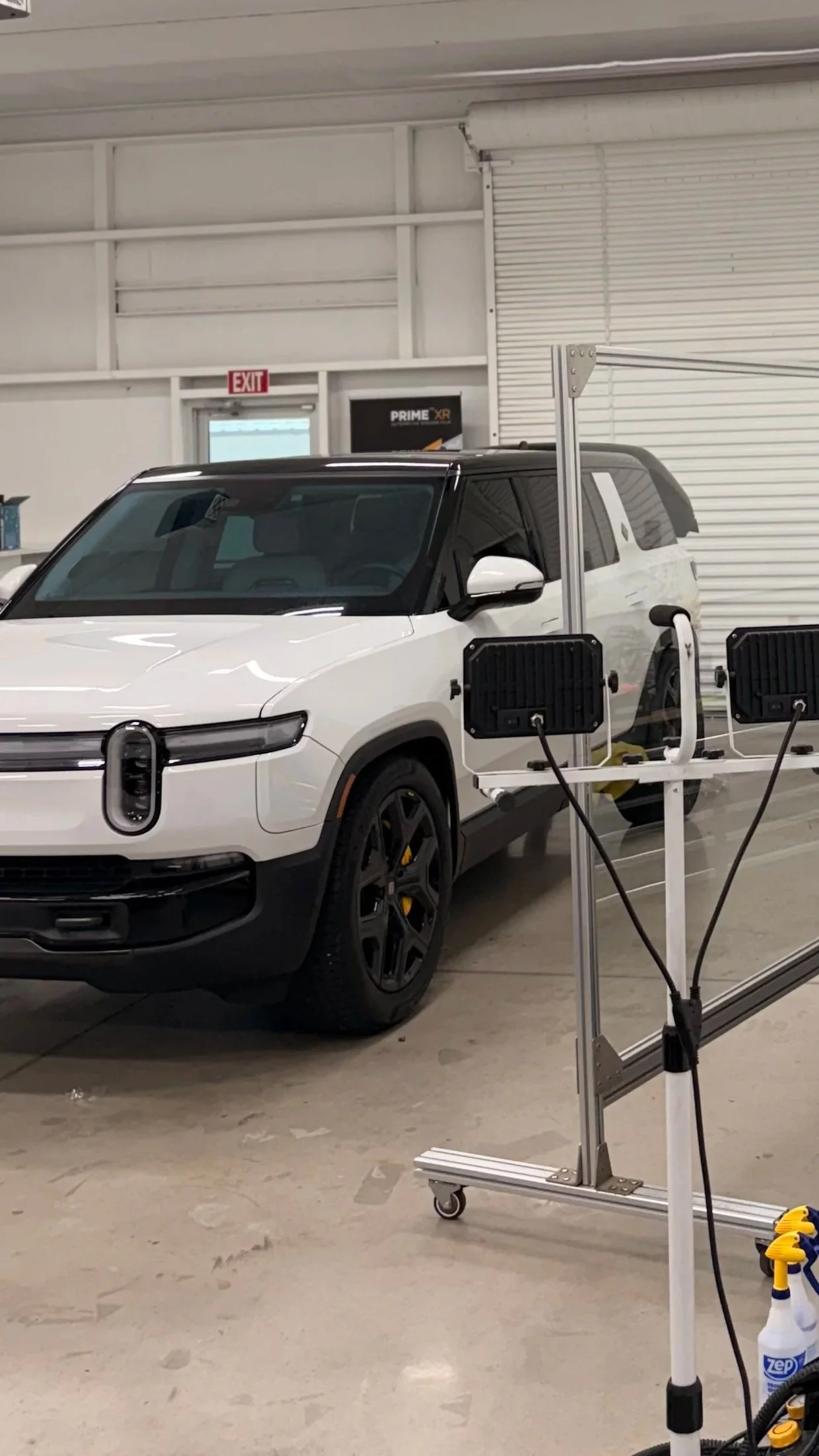 White electric vehicle inside a garage, connected to charging equipment. The garage has white walls, a steel roll-up door, and an exit sign above a door in the background.