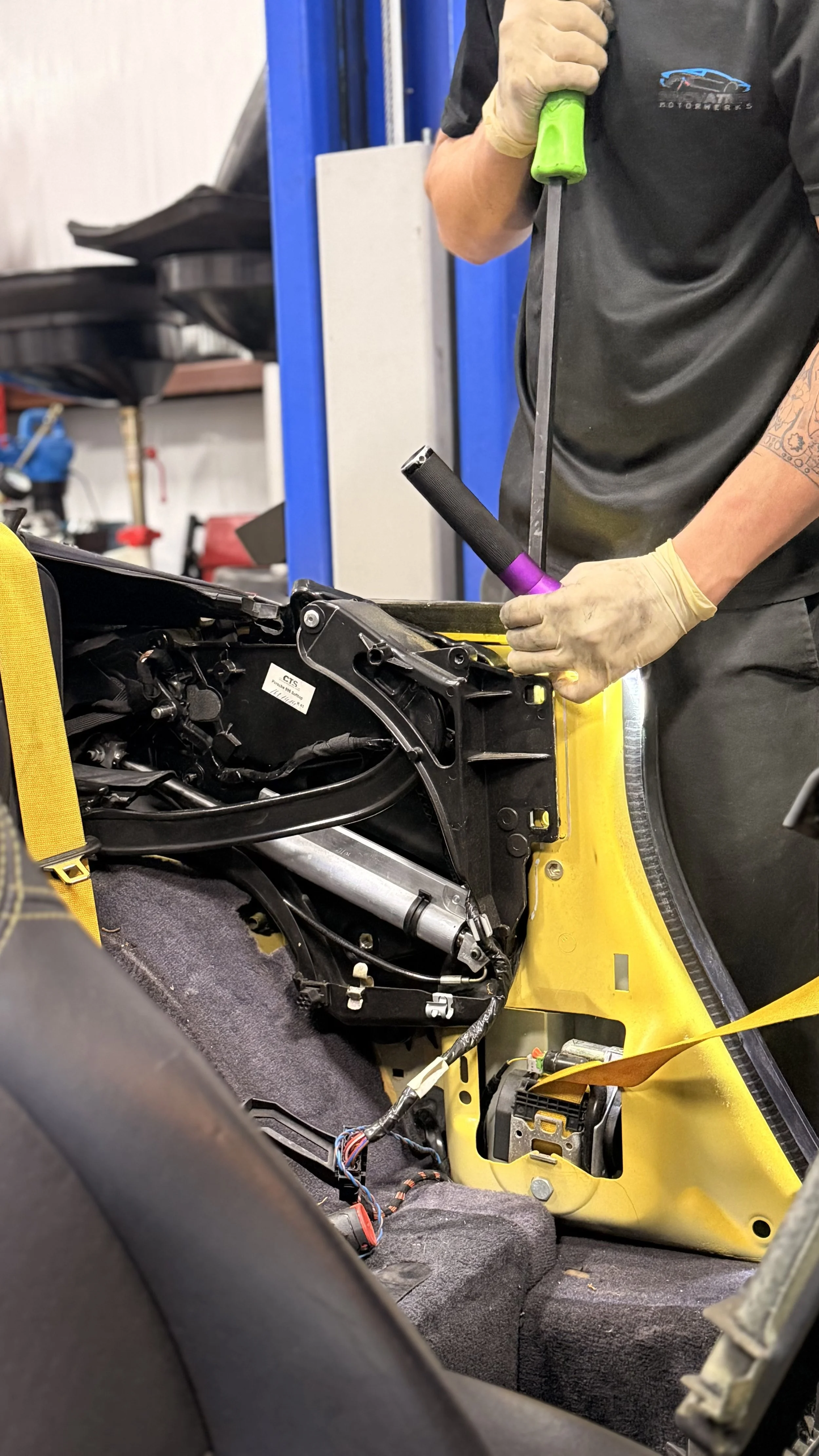 A mechanic working on a yellow motorcycle frame using a crowbar. The motorcycle's interior wiring and components are exposed, with the mechanic wearing gloves and a black shirt. The workshop background has tools and equipment.