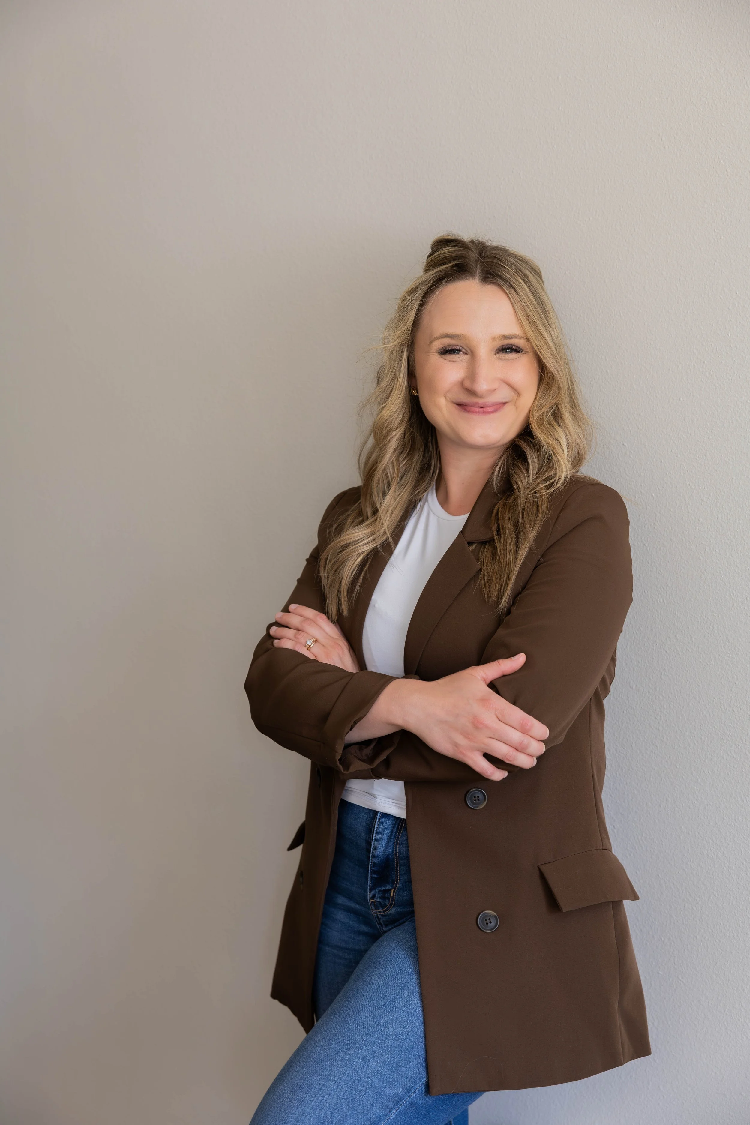 Portrait of a woman with blonde wavy hair wearing a brown blazer over a white top, crossed arms, standing against a plain beige wall, smiling.
