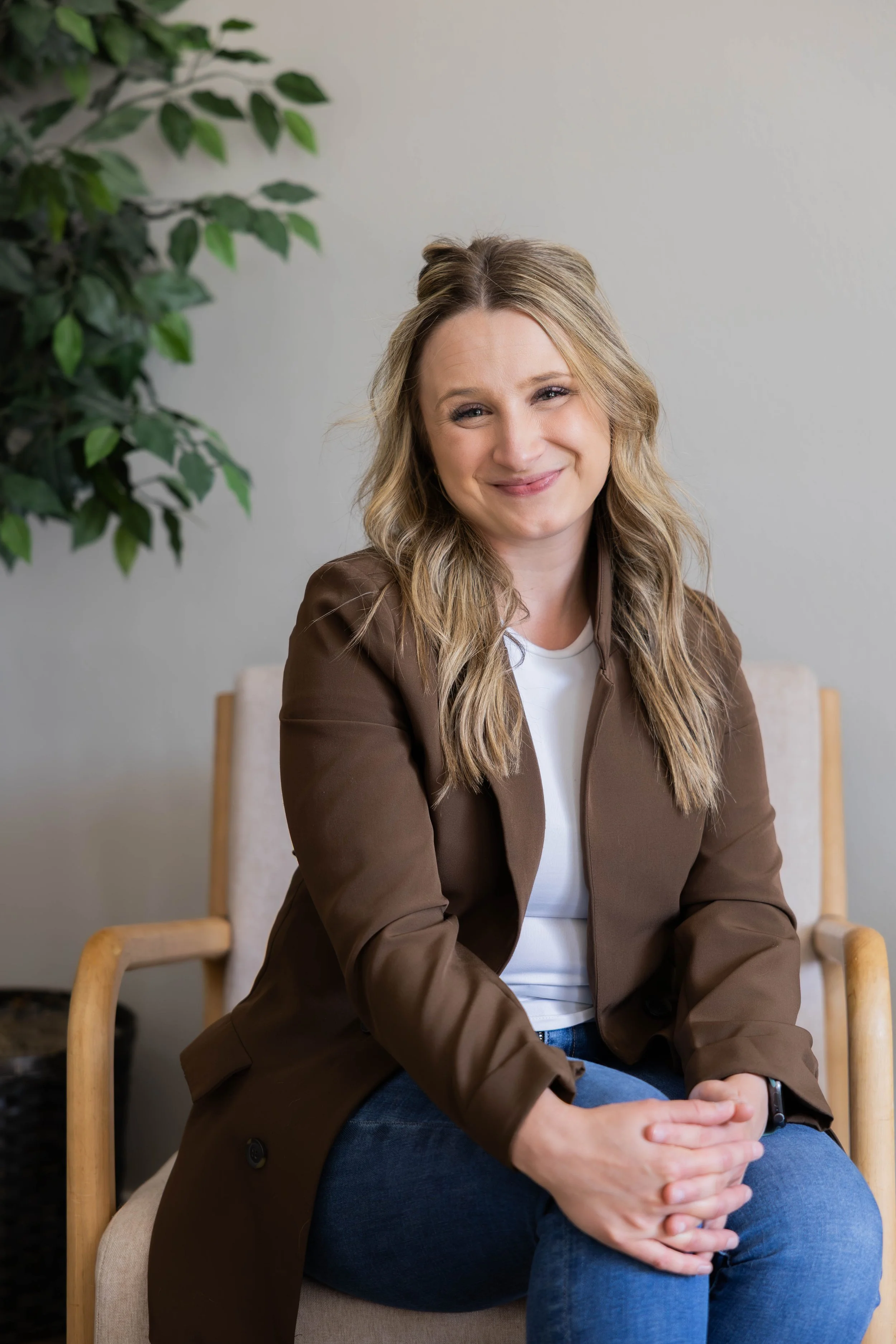 A woman with wavy blonde hair, smiling, wearing a white top and brown blazer, sitting on a beige chair with wooden armrests. A green leafy plant is partially visible on the left against a plain light-colored wall.