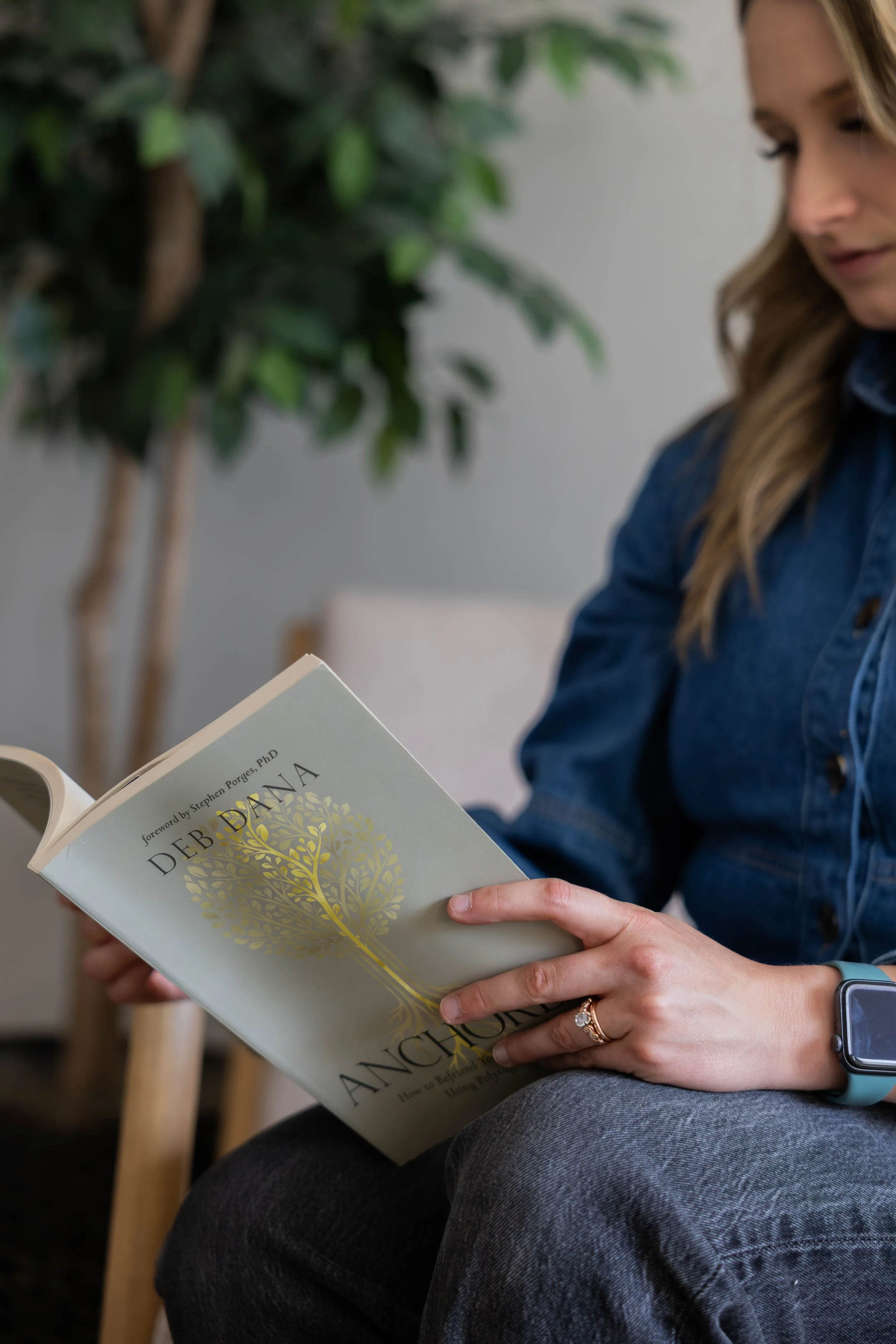 A woman sitting indoors reading the book "Angela" by Stephen Porges, PhD, wearing a denim jacket and a smartwatch.