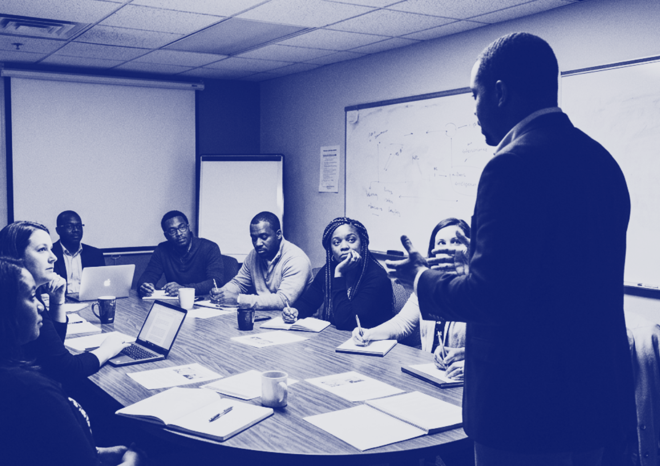 A group of diverse mental health professionals sitting around a conference table in a meeting room, listening to a man standing and speaking, with a whiteboard, flip chart, and documents on the table.