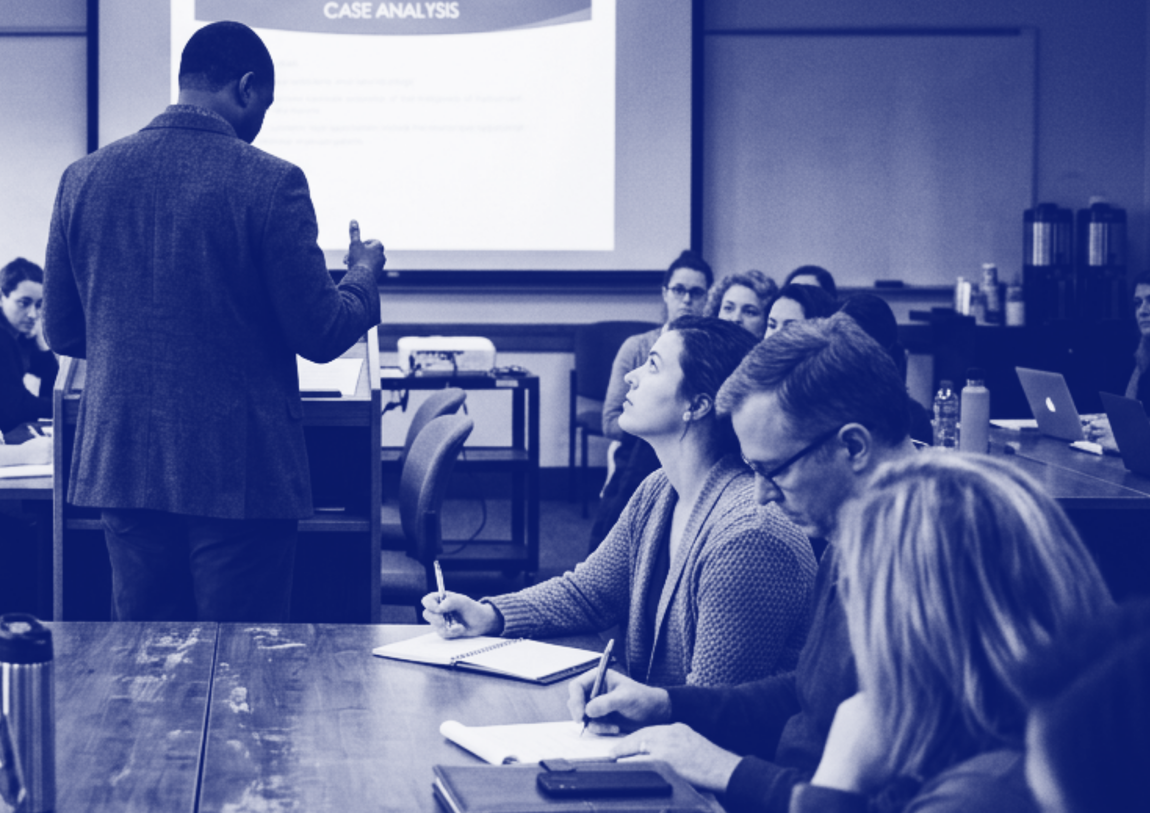 A mental health professional giving a presentation in a classroom with students seated at tables, some taking notes, with a large screen displaying 'Case Analysis' behind him.