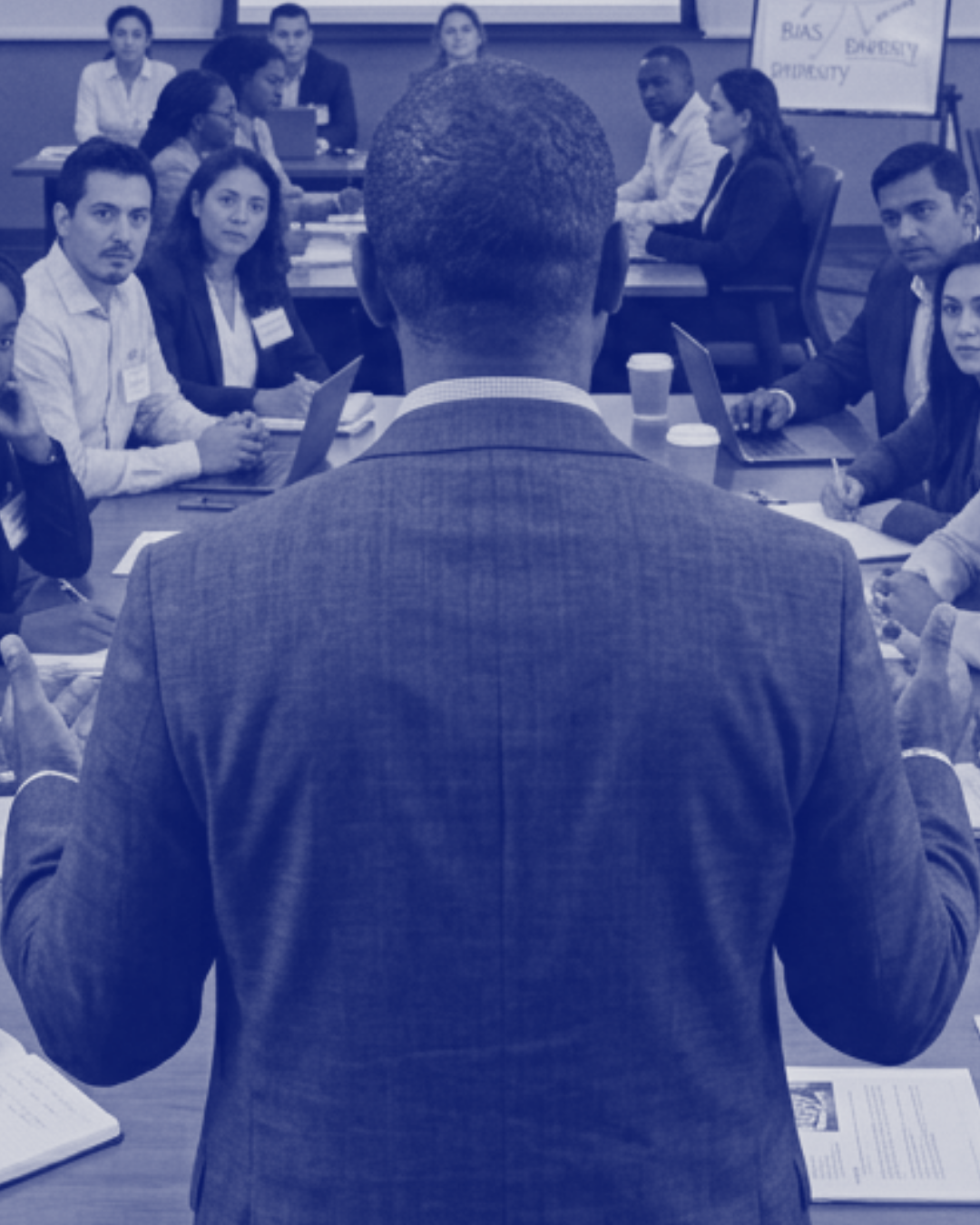 A mental health professional in a suit is giving a presentation or speech to a group of professional people seated around a conference table in a business meeting or workshop.