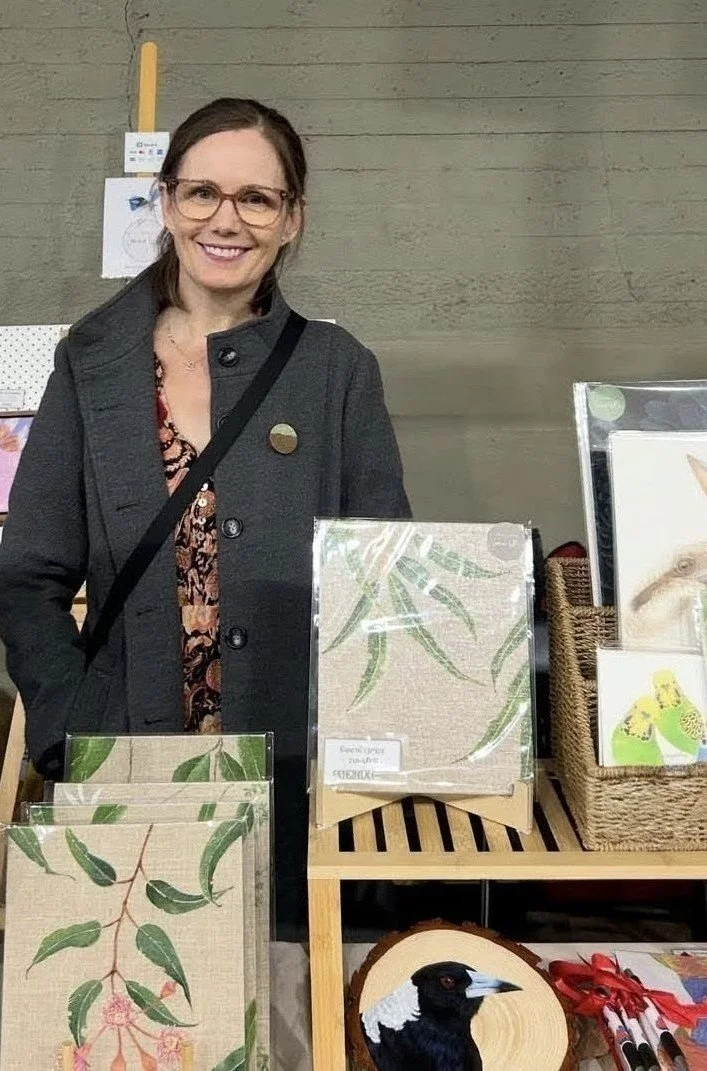Woman standing behind a display table with botanical artwork, art prints, and a wood plaque featuring a bird portrait.