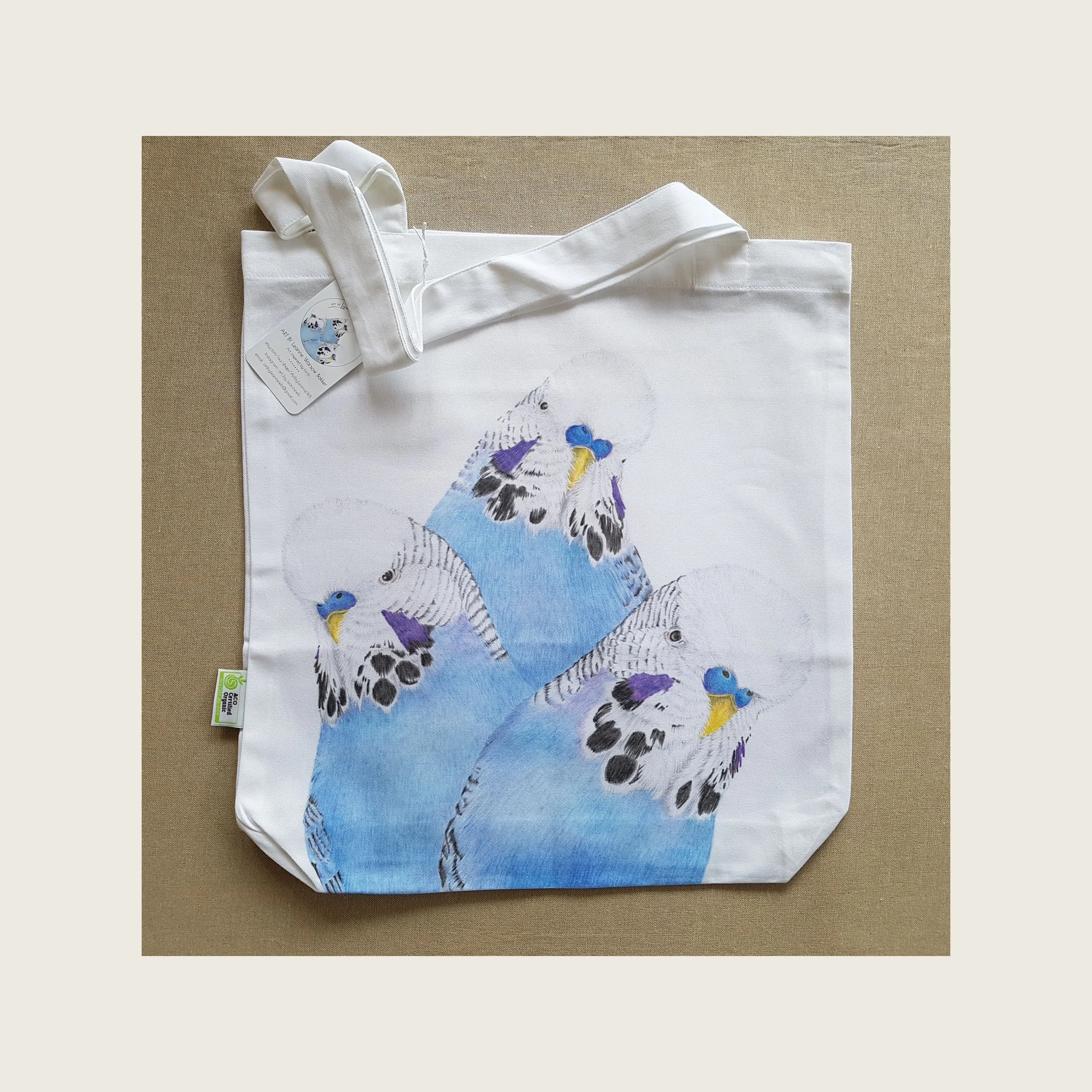 A white tote bag with an illustration of three birds on it.  The bird is a blue budgie in three different poses.