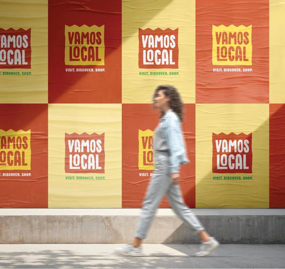 A woman walking past a large, colorful poster wall with repeated 'VAMOS LOCAL' slogans and the subtitle 'visit. discover. shop.' in green.