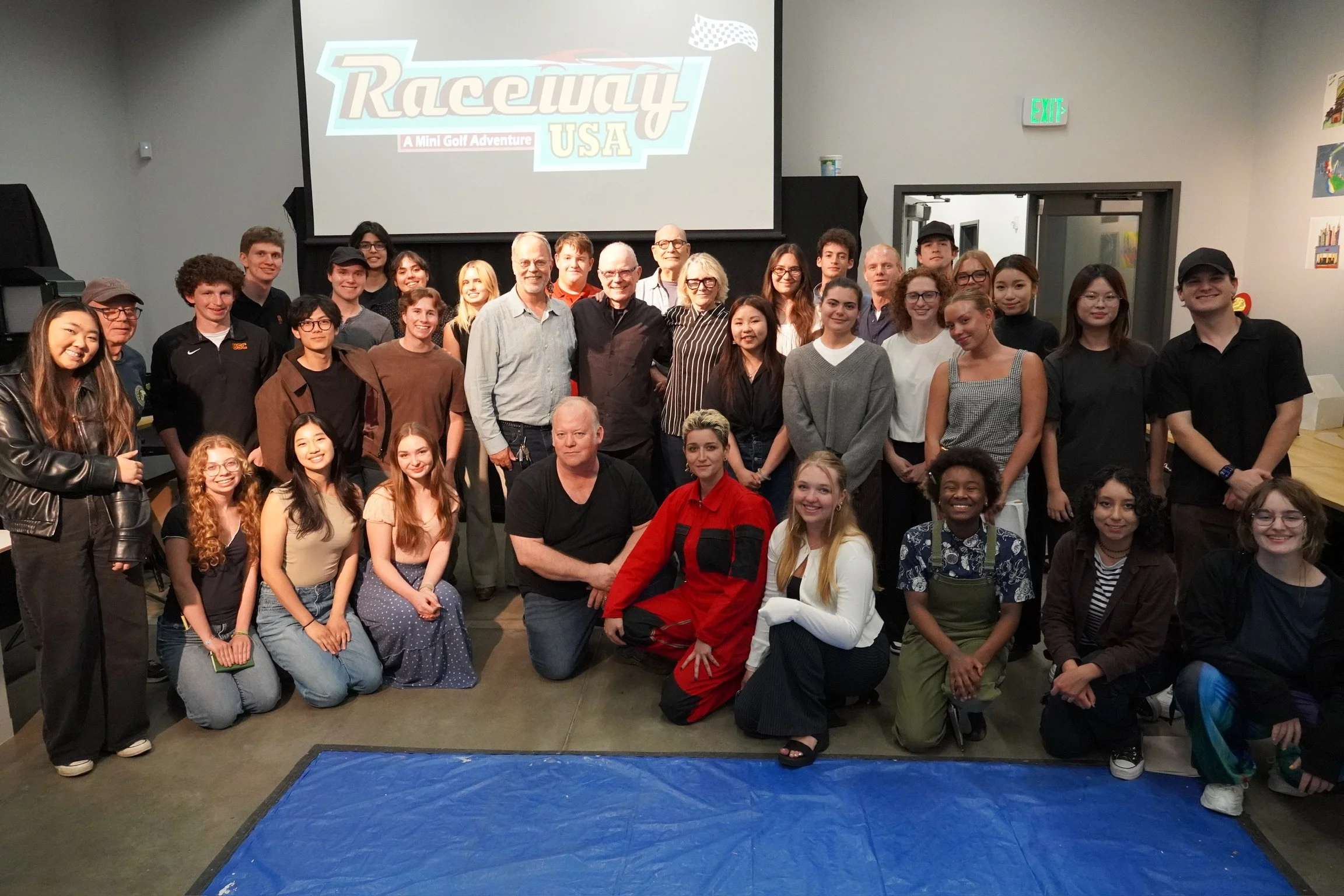 A group of diverse people, including adults and children, posing for a photo in front of a large screen displaying the logo for 'Racceway USA: A Mini Golf Adventure'. They are indoors, some are smiling, and a blue mat is visible on the floor in the foreground.