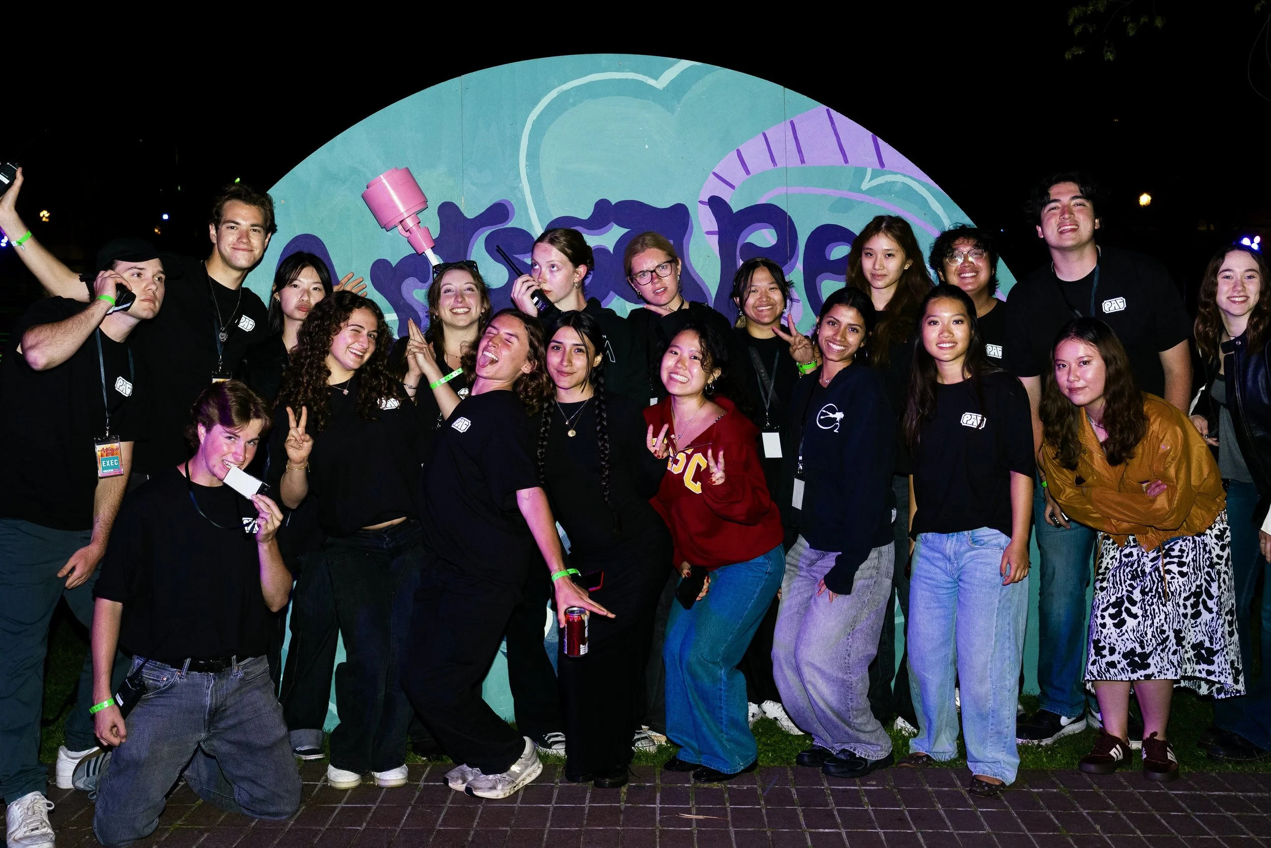 Group of diverse young people smiling and posing together at night in front of a colorful mural with abstract shapes and a smiling face.
