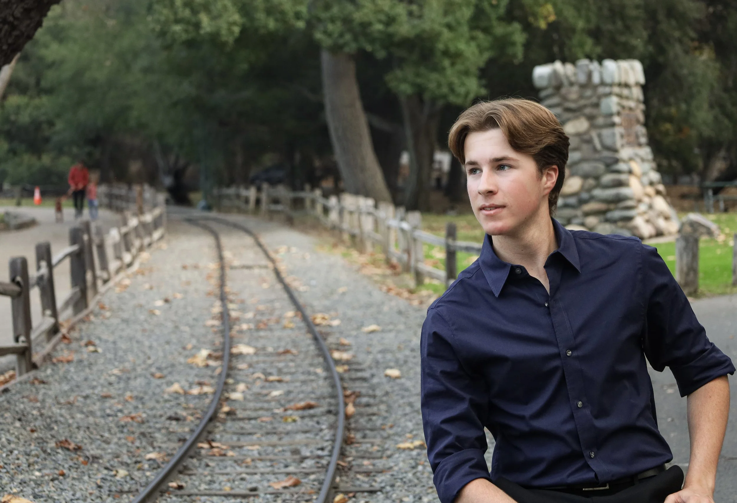 A young man with brown hair wearing a dark blue button-up shirt standing outdoors on a cloudy day near a gravel train track. There are trees and a stone structure in the background, and two people walking a dog in the distance.