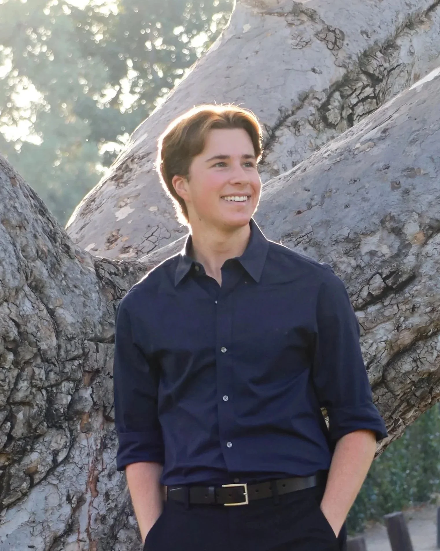 A young man with brown hair, wearing a black shirt and black belt, is smiling and leaning against a large tree with textured bark. The background features blurred sunlight and greenery.