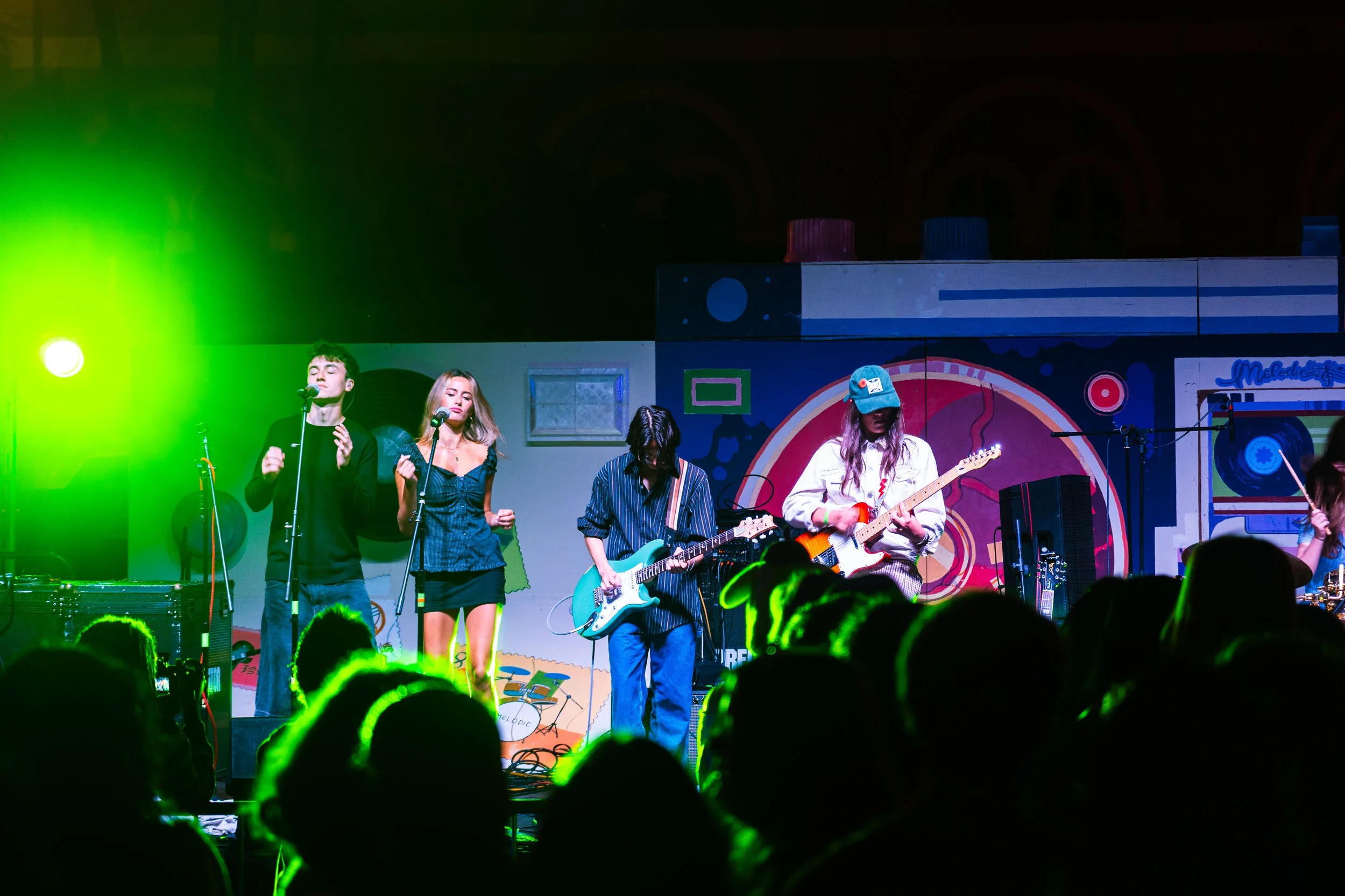 A band performs on stage with colorful background. Four musicians are visible, including two females singing into microphones and playing guitars, and two males playing electric guitars. Audience members' heads are seen in the foreground, illuminated with green lighting.
