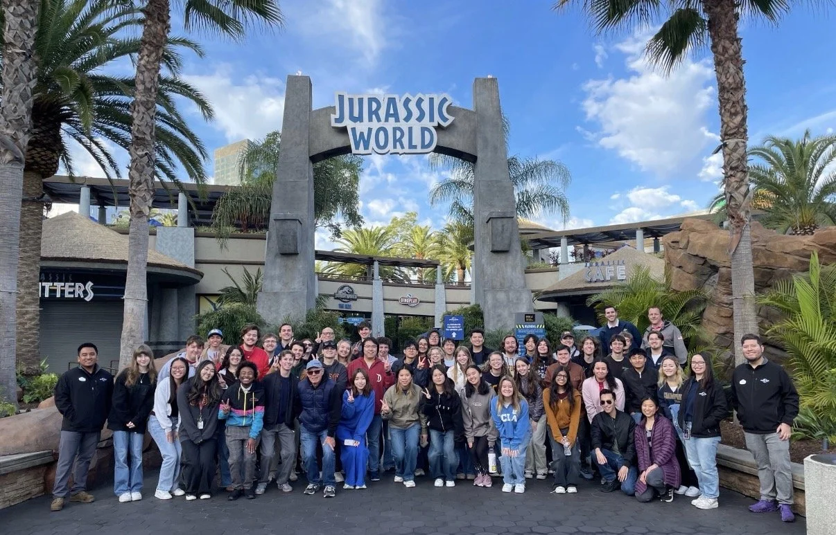 A large group of diverse people posing in front of the Jurassic World sign at an amusement park entrance on a sunny day, with palm trees and park buildings in the background.