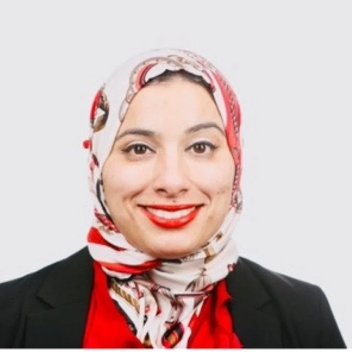 A woman wearing a black blazer and red blouse, with a colorful patterned headscarf, smiling against a plain white background.