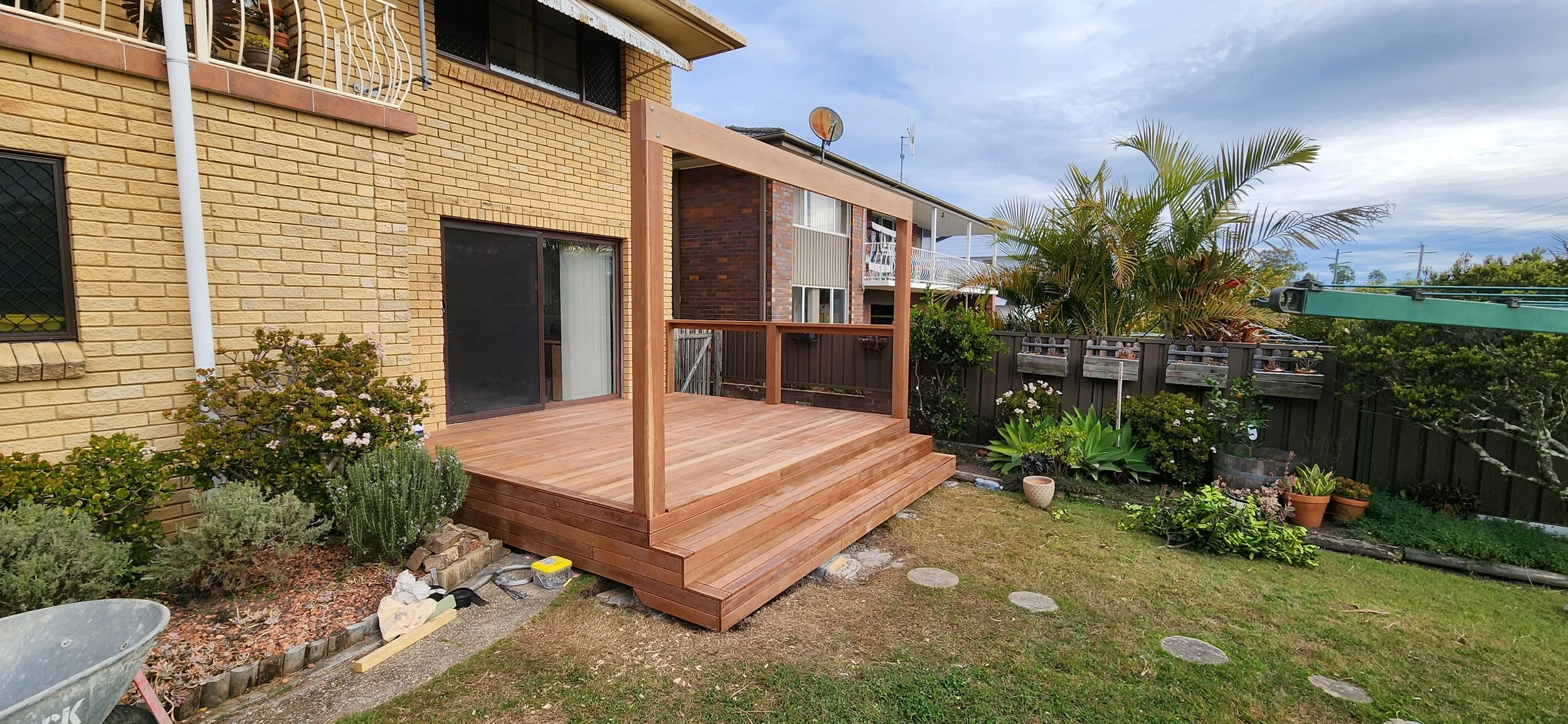 A backyard with a newly built wooden deck attached to a yellow brick house. There are plants and bushes around the deck, a black fence, and a sky with some clouds. The deck has a railing and steps leading down to the grass yard.