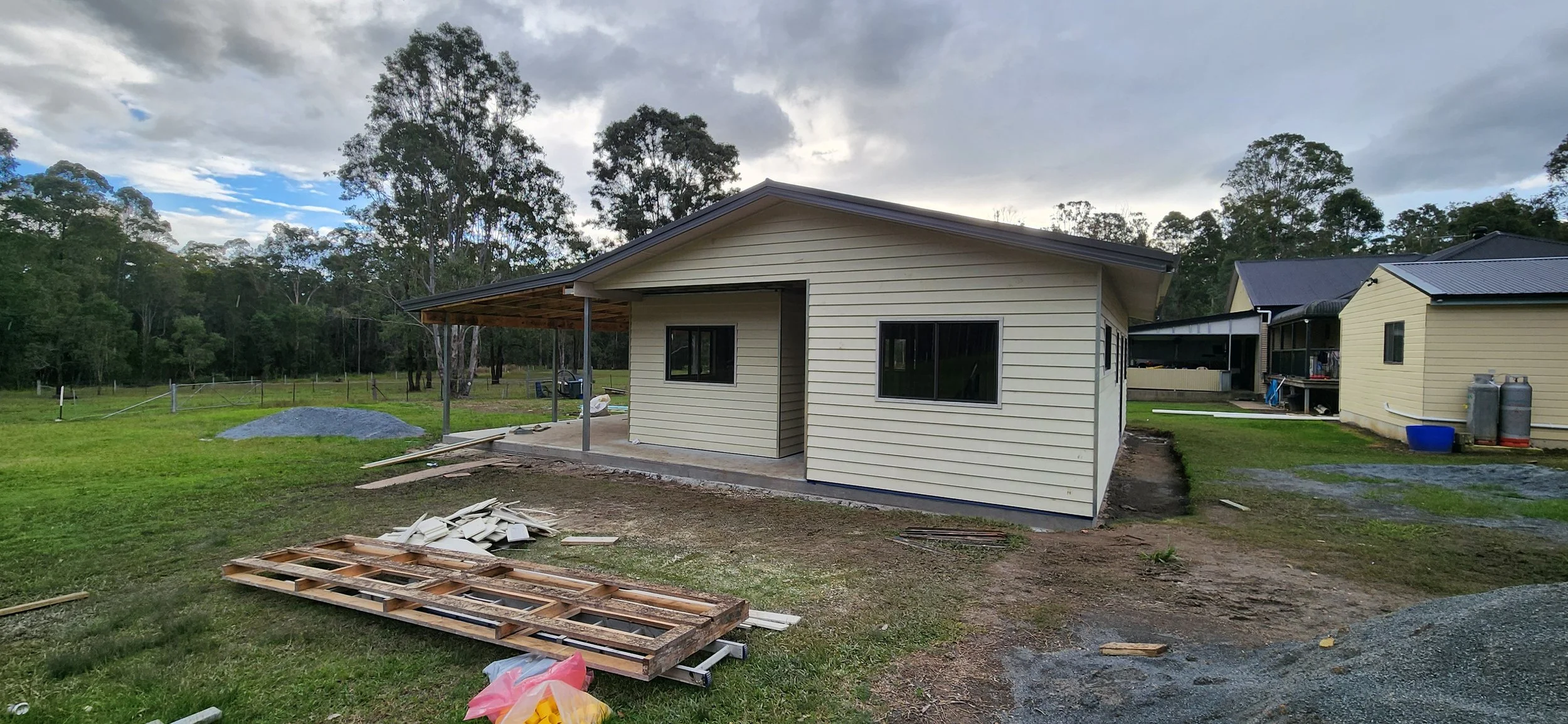 A house under construction in a grassy area with trees in the background, showing exterior walls and windows, with construction materials and tools scattered around.