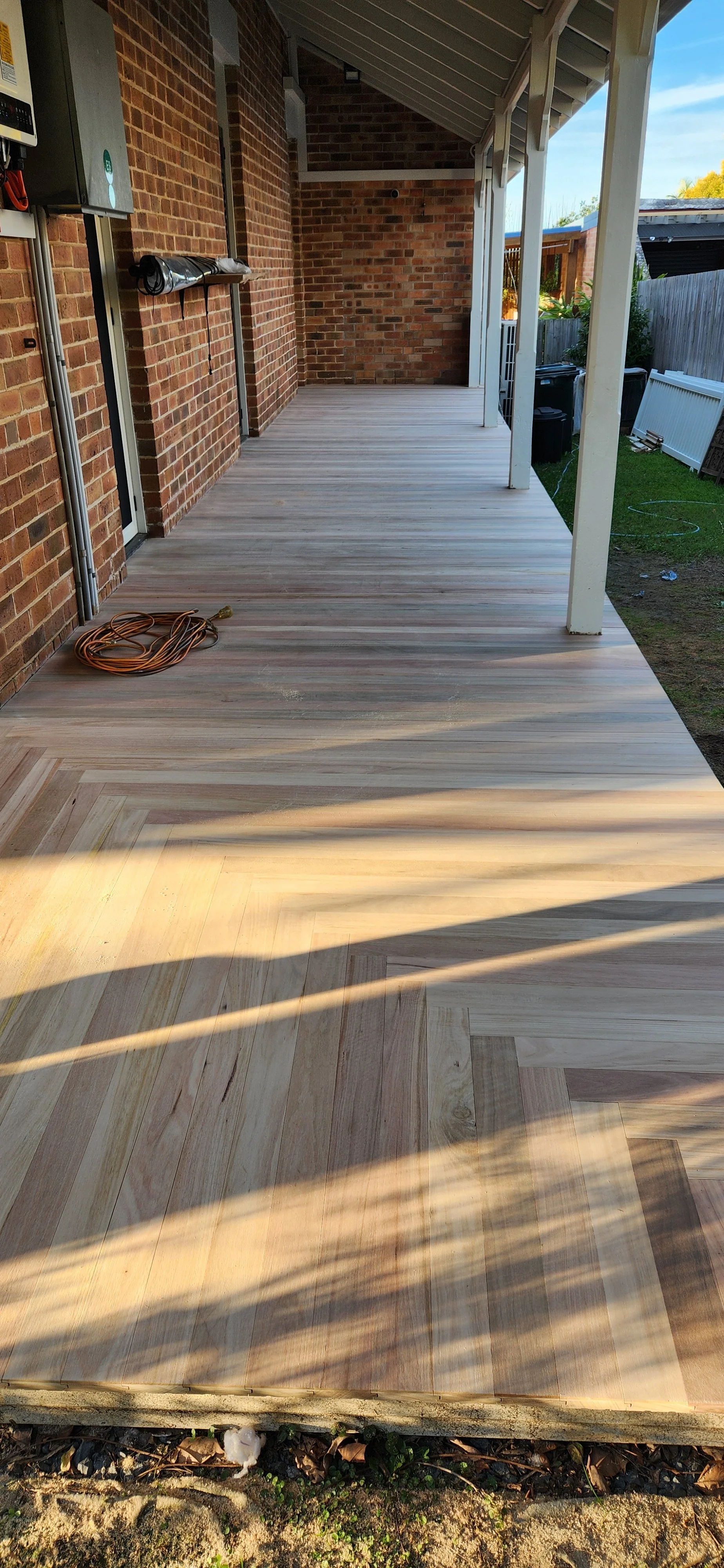 Newly installed wooden outdoor deck attached to a brick house with a covered porch, electrical cords on the deck, and a grassy yard with a fence and trees in the background.