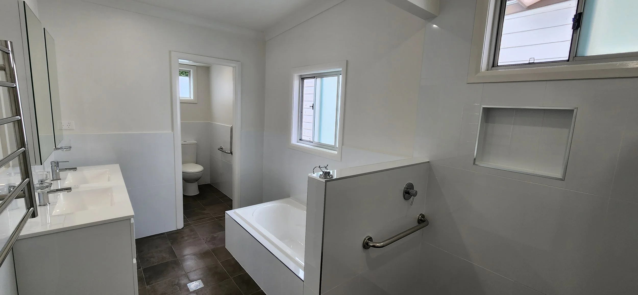 Modern bathroom with white walls, a bathtub, a toilet, two windows, and a vanity with a double sink.