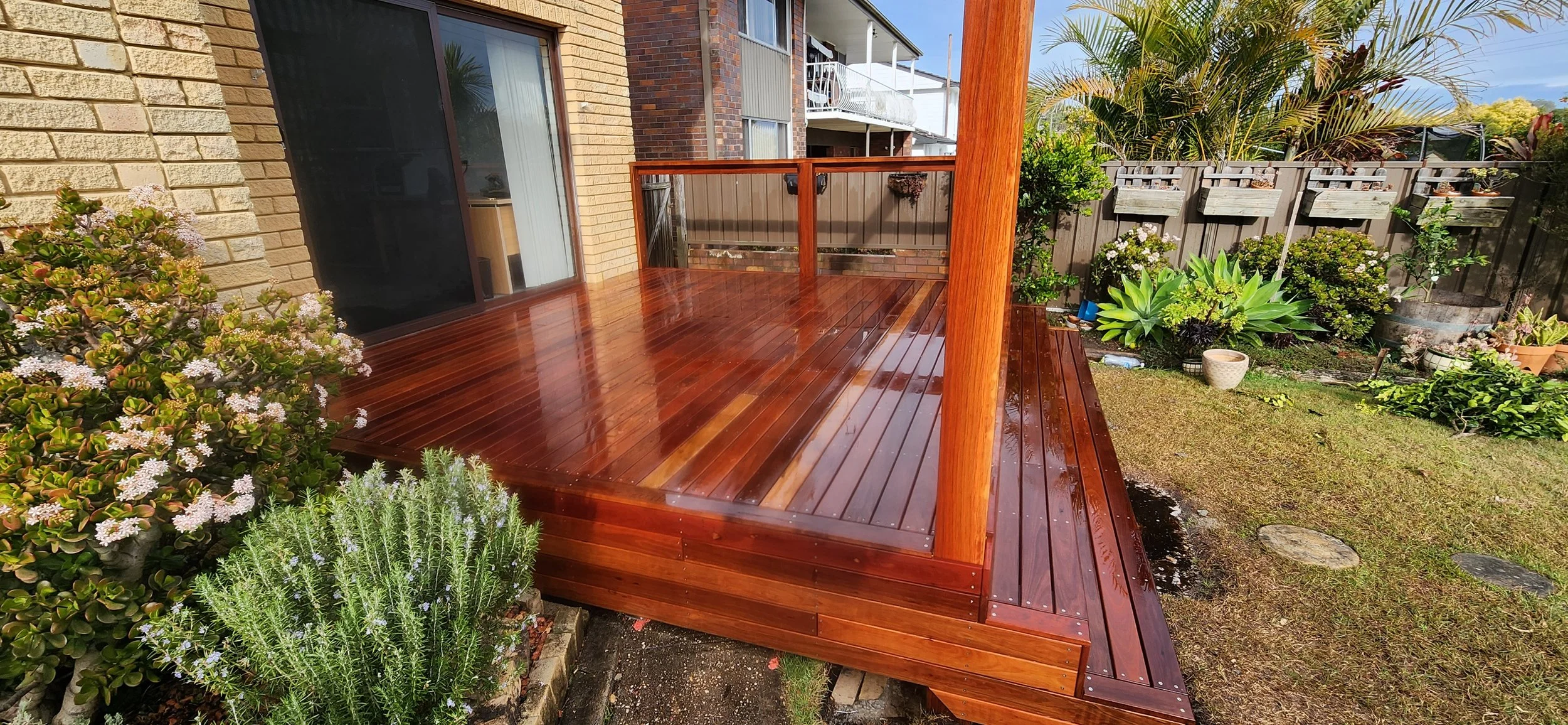 A newly polished wooden backyard deck attached to a house, surrounded by a lush garden with various plants and flowers, a brick wall, and a wooden fence.