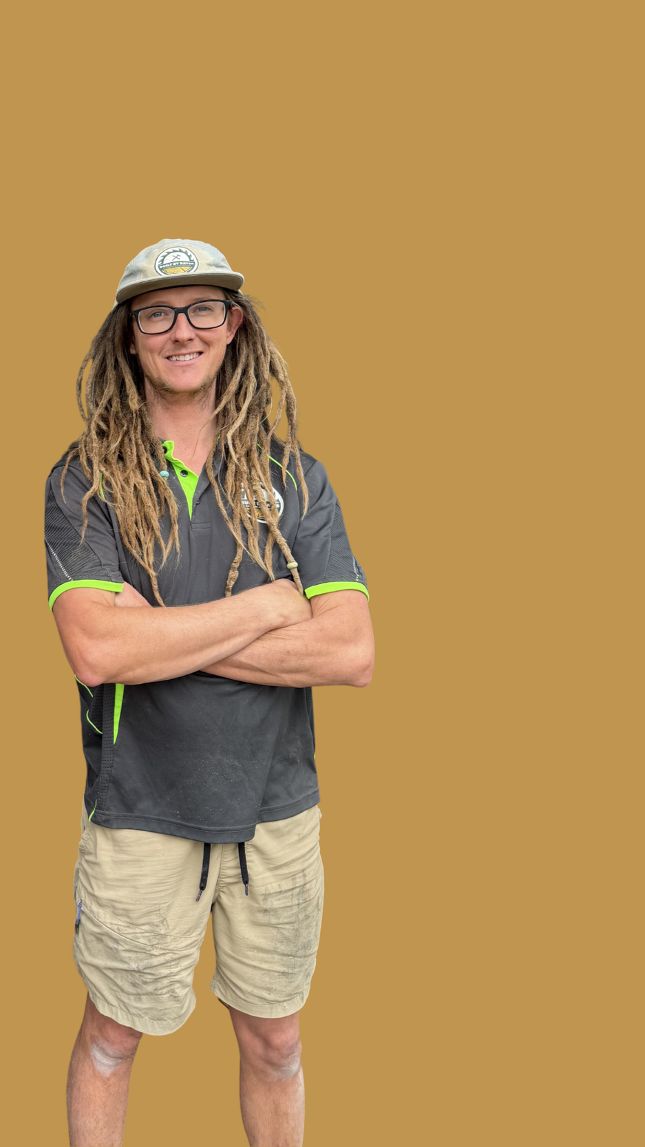 A man with dreadlocks wearing glasses, a hat, a dark gray polo shirt with bright green accents, and khaki shorts, standing with arms crossed against a plain, solid tan background.