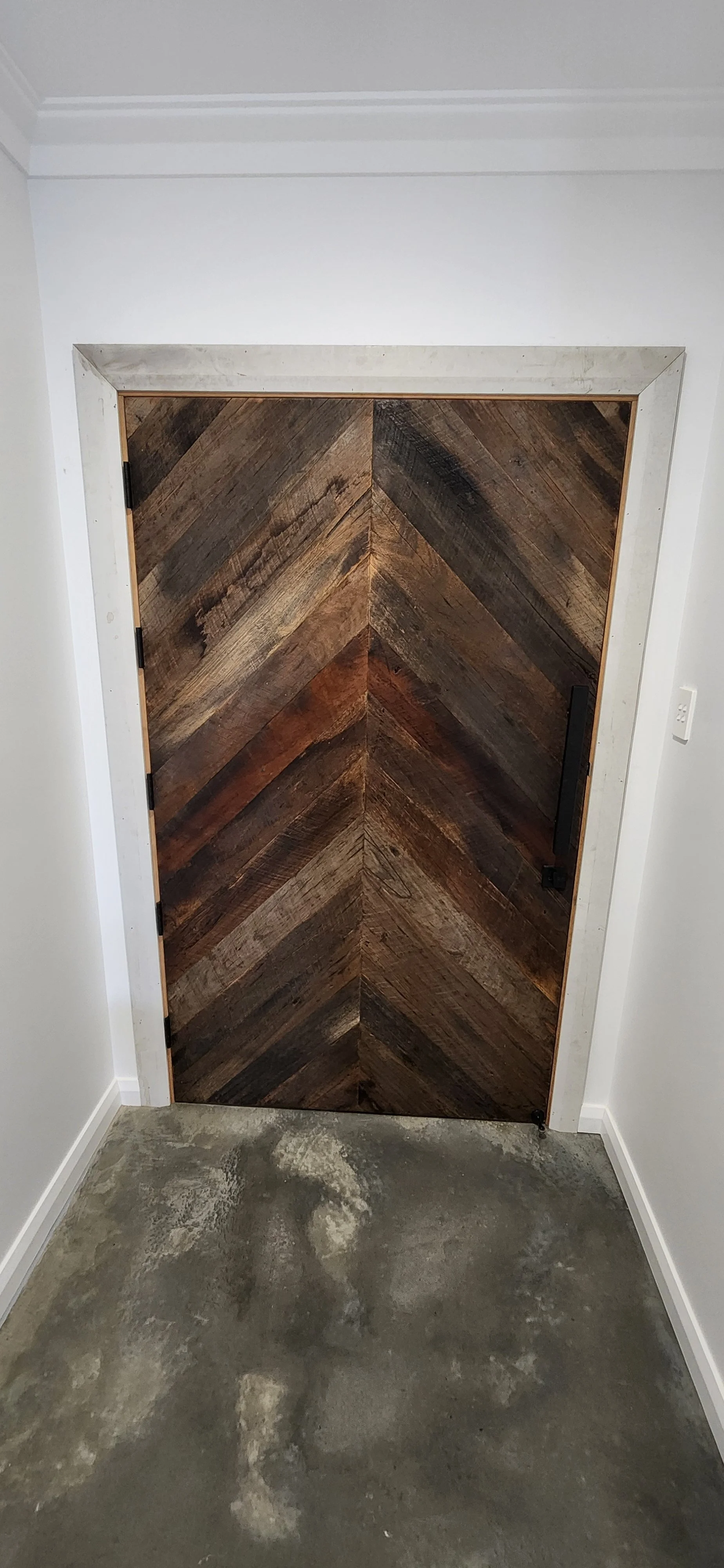 A wooden door with a chevron pattern mounted in a white wall, with a concrete floor in front.