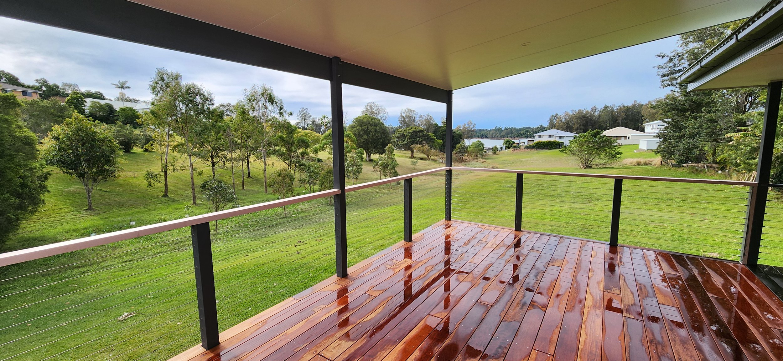 A wooden balcony overlooking a lush green landscape with trees and houses in the distance, under a partly cloudy sky.