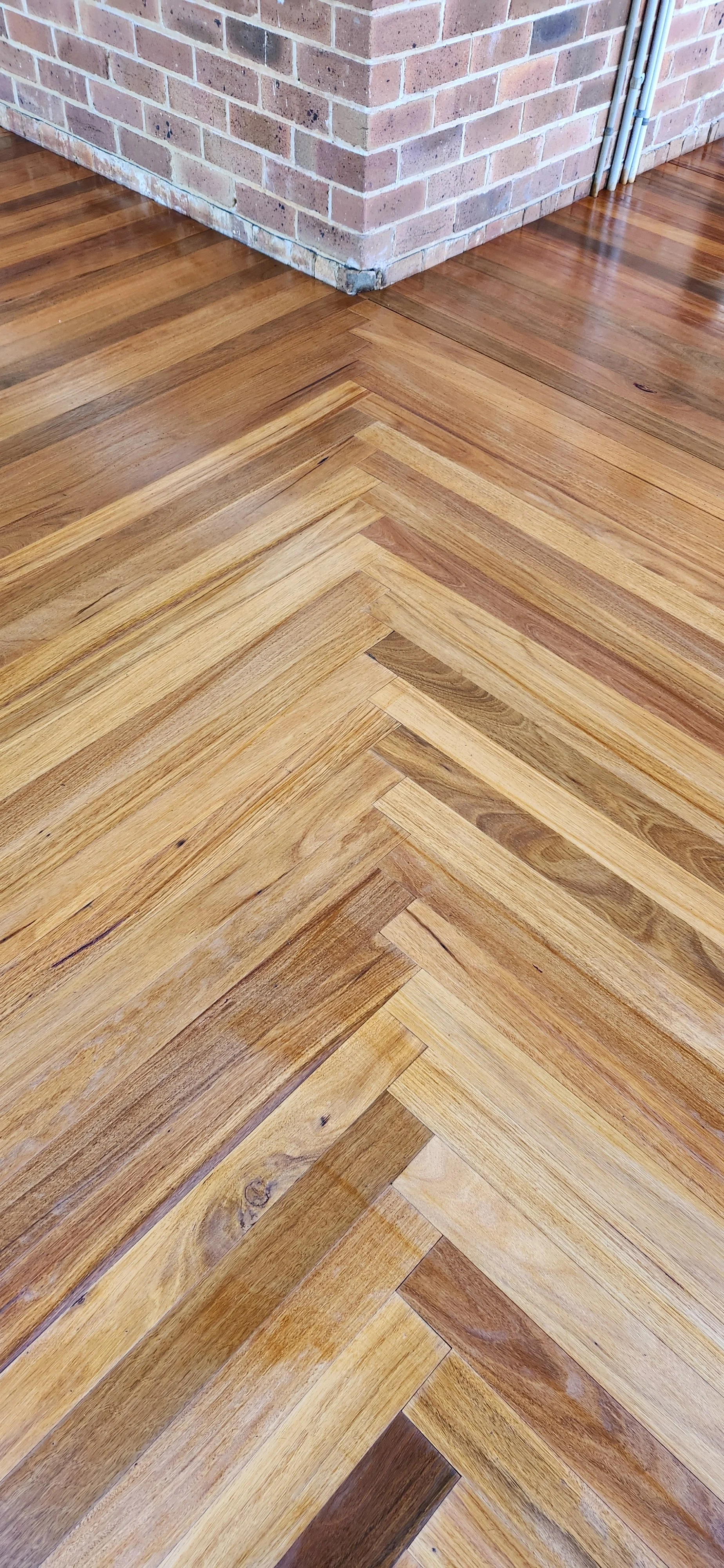 Wooden parquet flooring arranged in a herringbone pattern next to a brick wall with visible corner and piping.