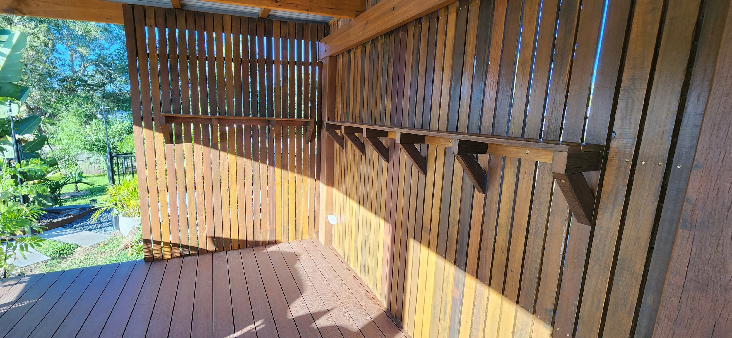 View of a wooden balcony with a railing, surrounded by green trees and plants, with a glimpse of the backyard.