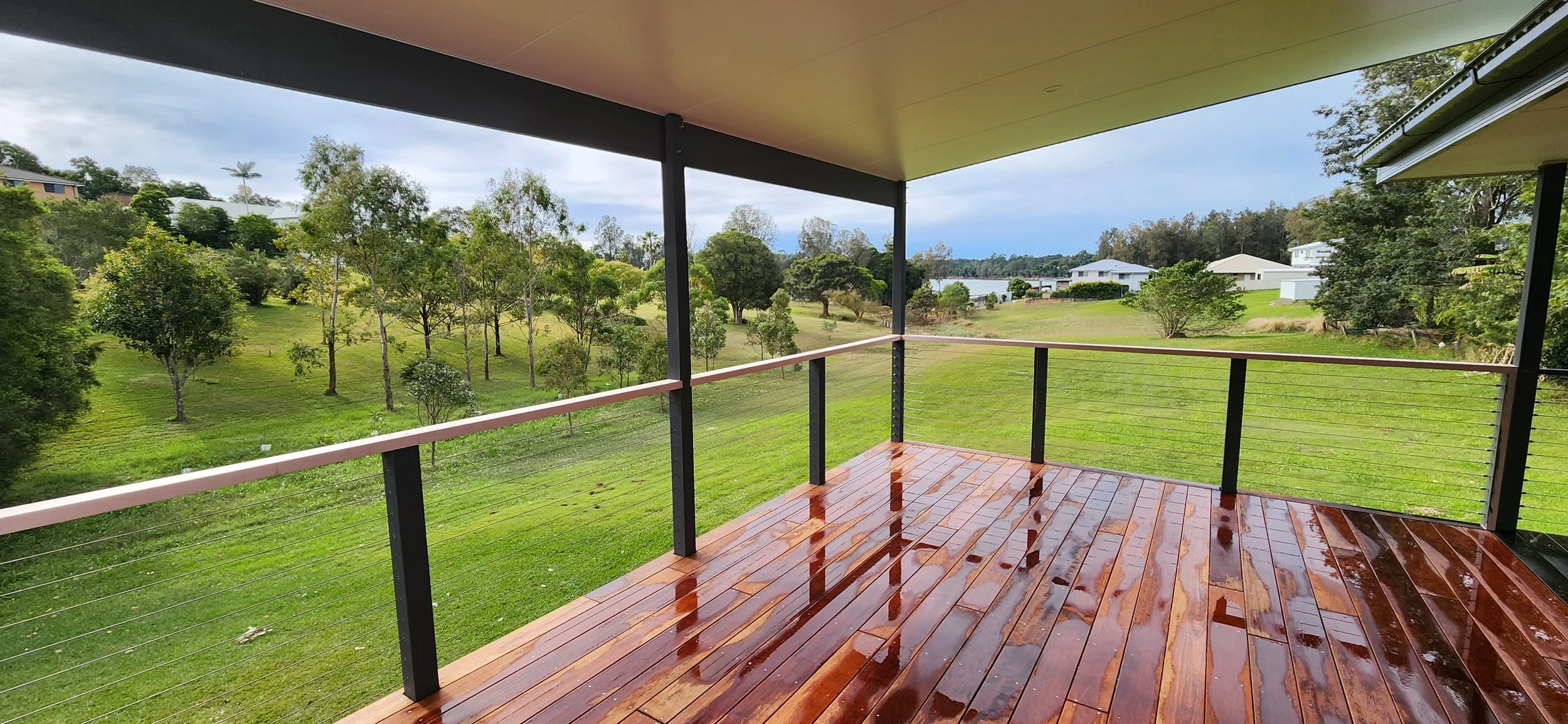 View from a covered balcony with wet wooden floor overlooking lush green yard with trees and houses in the distance under a cloudy sky.