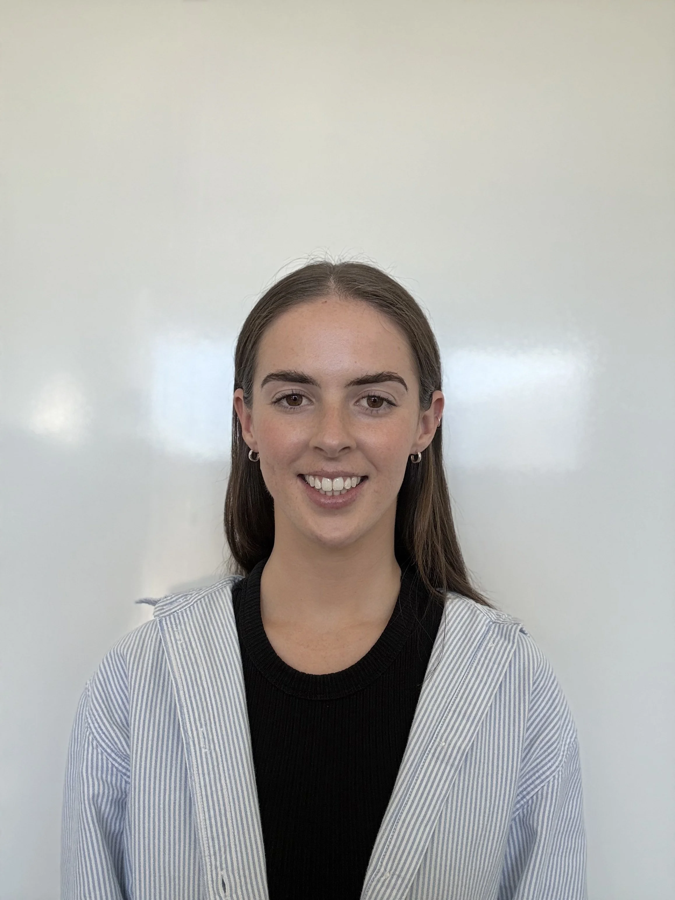A young woman with shoulder-length brown hair and earrings, smiling, wearing a black top and a light blue striped blazer, standing in front of a plain white wall.