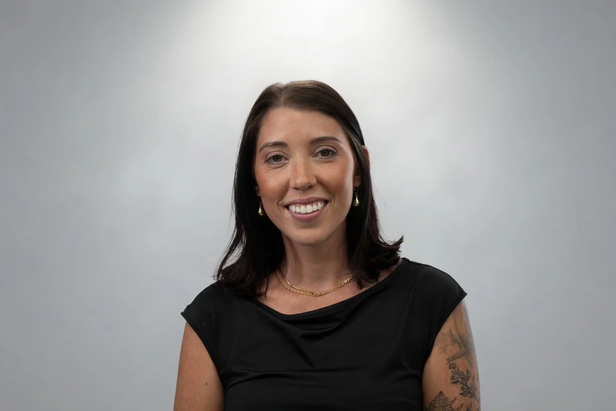 A woman with shoulder-length dark hair smiling at the camera, wearing a black top, gold jewelry, and earrings, standing against a plain light-colored background.