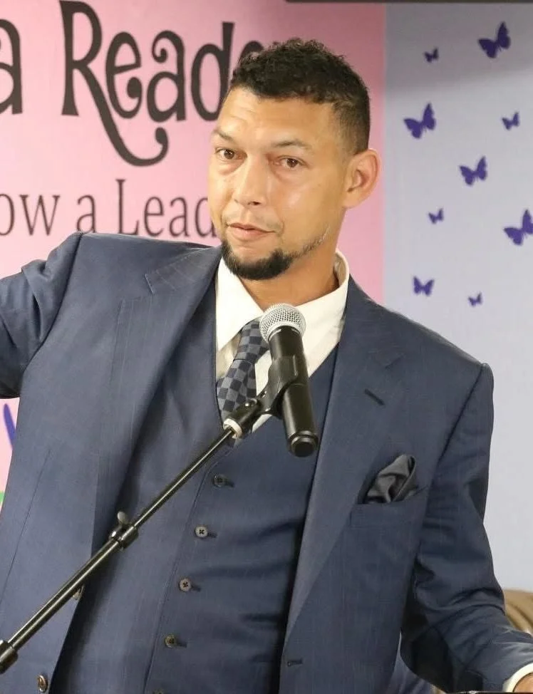 Man in a dark blue suit and white shirt speaking at a microphone in front of a pink and purple background with butterflies and text