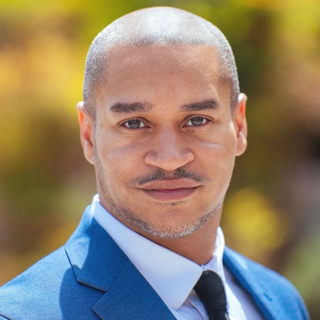 Close-up portrait of a man with a shaved head and goatee, wearing a blue suit and a black tie, outdoors with blurred yellow and green foliage in the background.