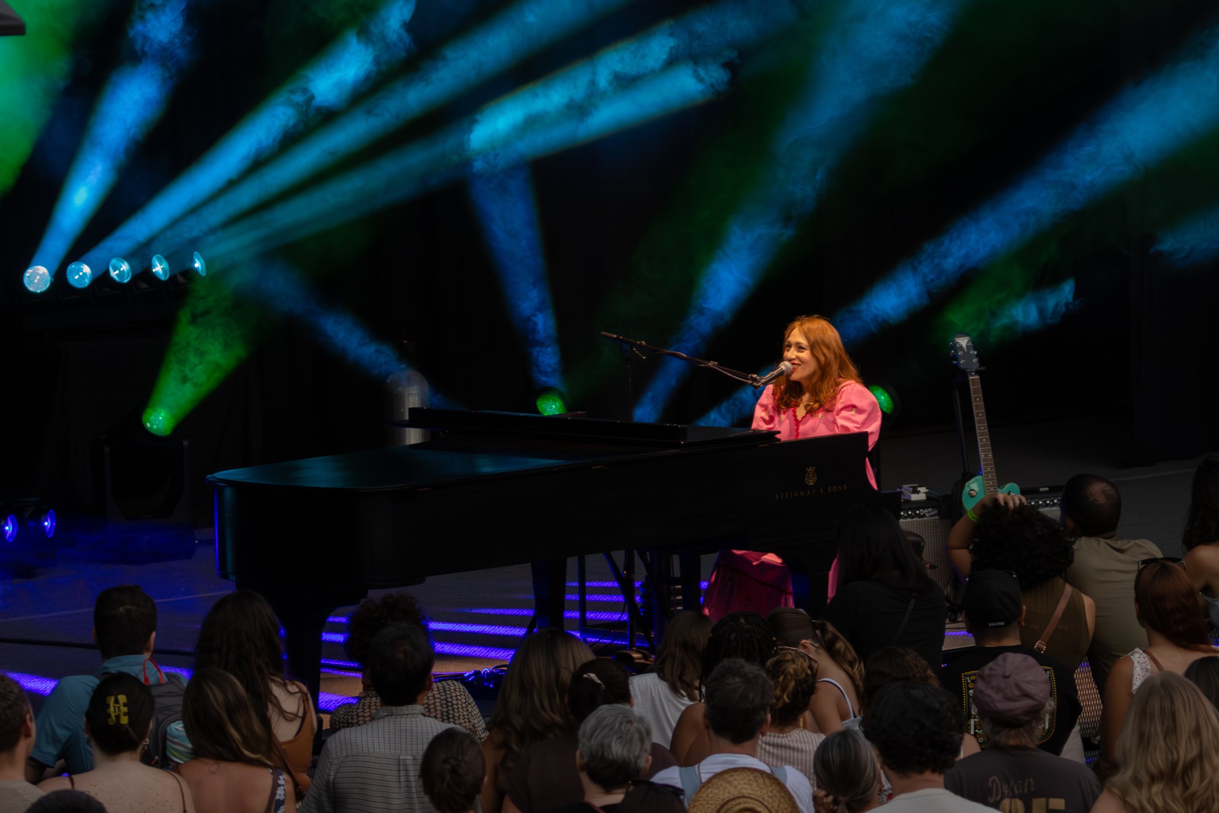 A woman with red hair wearing a pink dress playing a black grand piano on stage, smiling at the audience. The stage features colorful lighting effects with green, blue, and purple beams. Several children are seated on the floor in front of the stage,